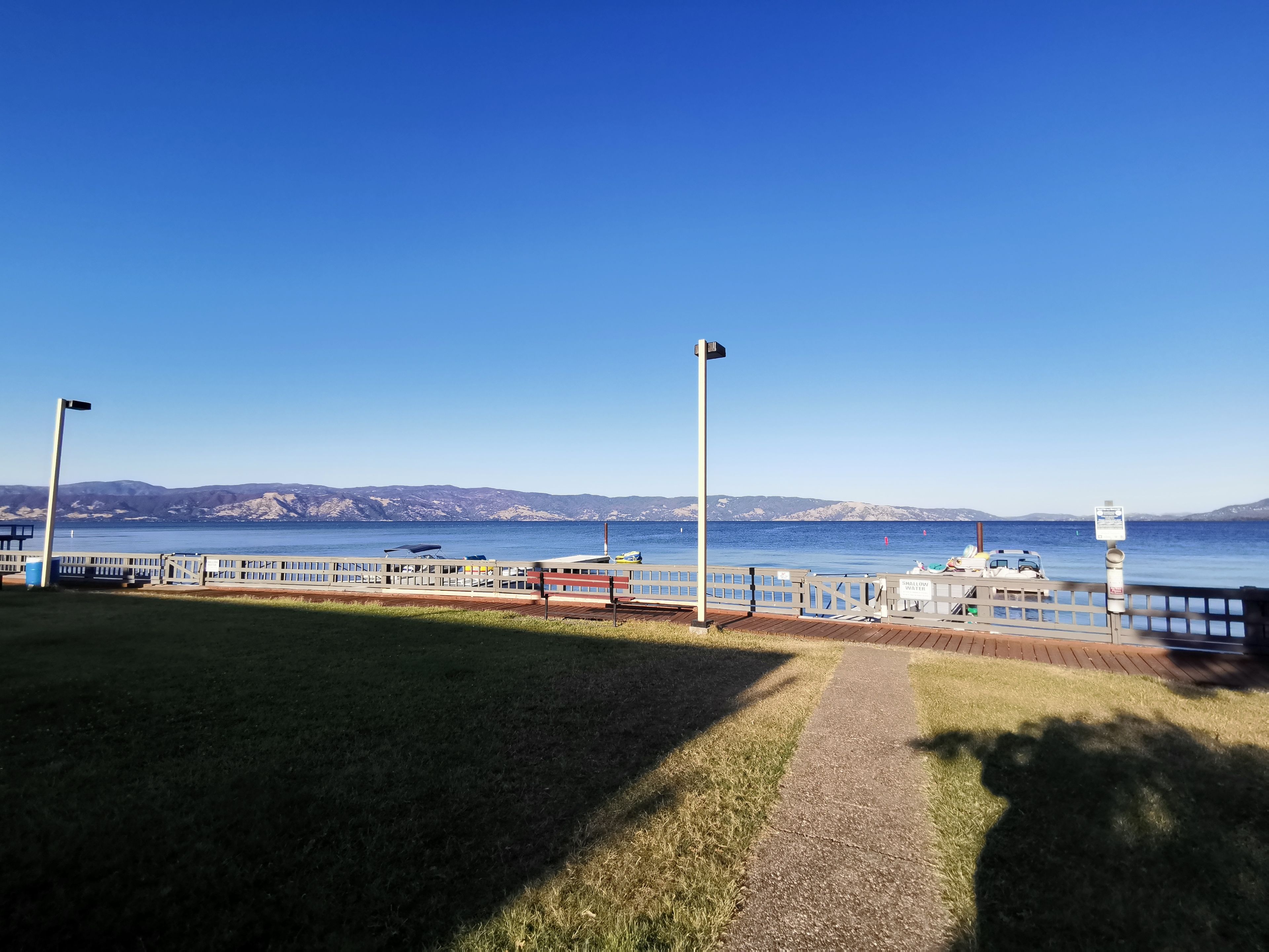 Freie Sicht auf ein Ufergebiet mit einem Weg, der zu einem Steg führt, einer Bank und einem Schild, das auf flaches Wasser hinweist.