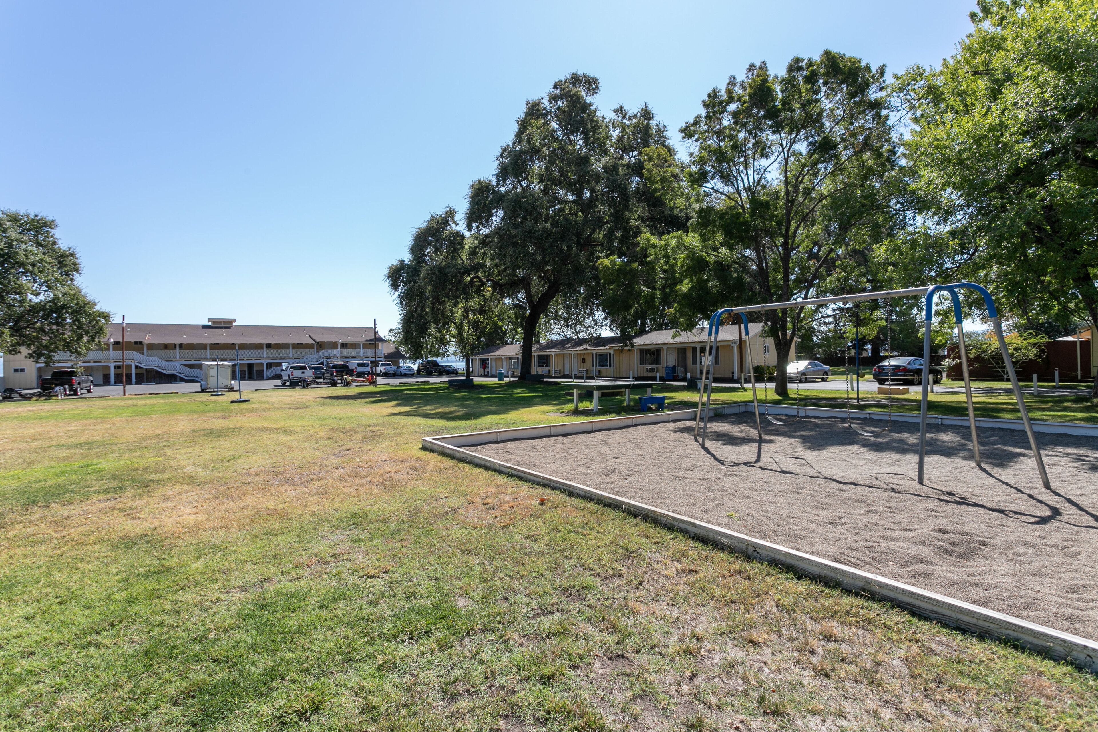 A playground with swings and a sandbox, surrounded by trees and a building with multiple units.