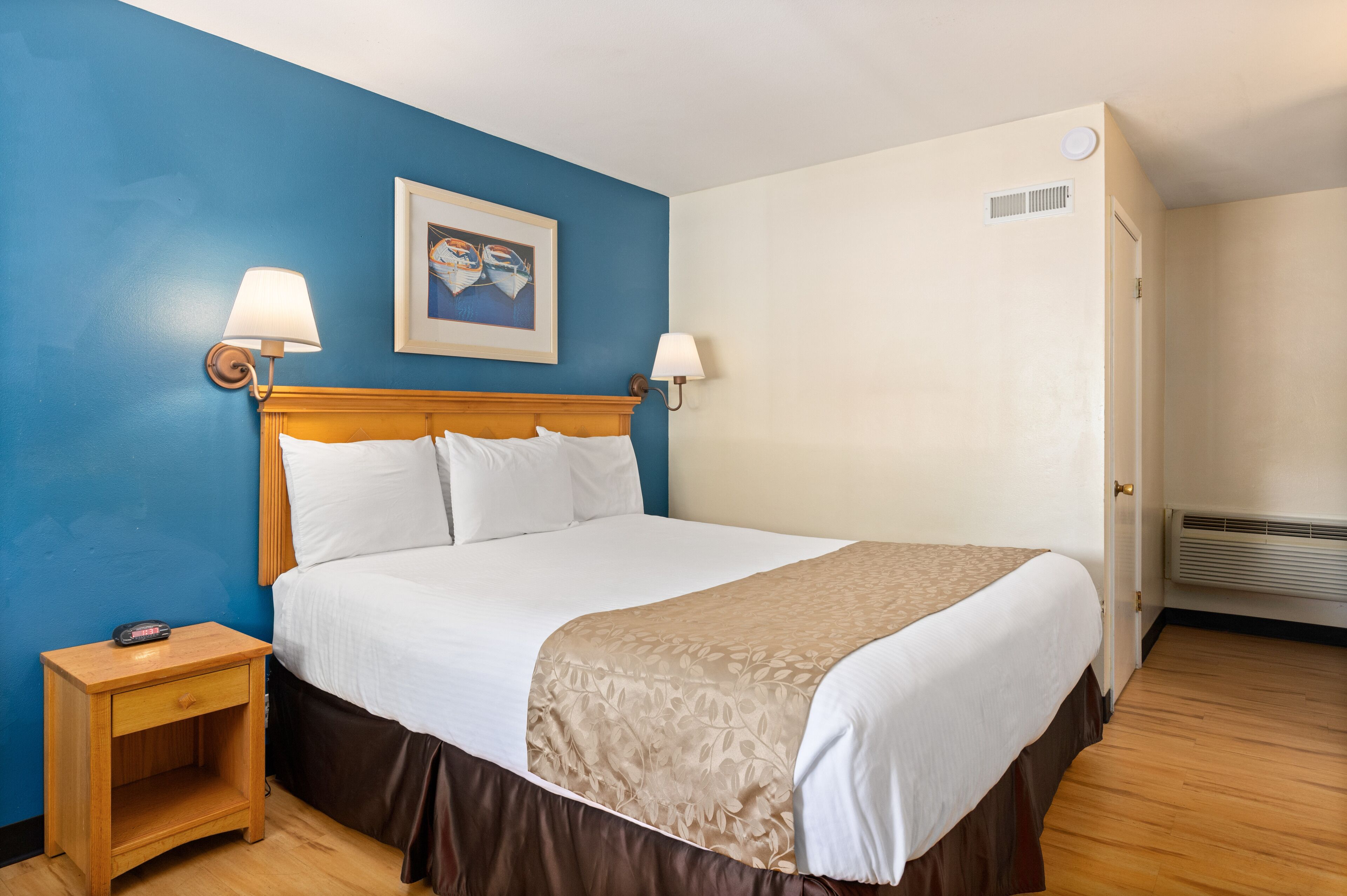 A hotel room with a bed, two bedside lamps, a nightstand, a framed picture on the wall, and a door with a vent.