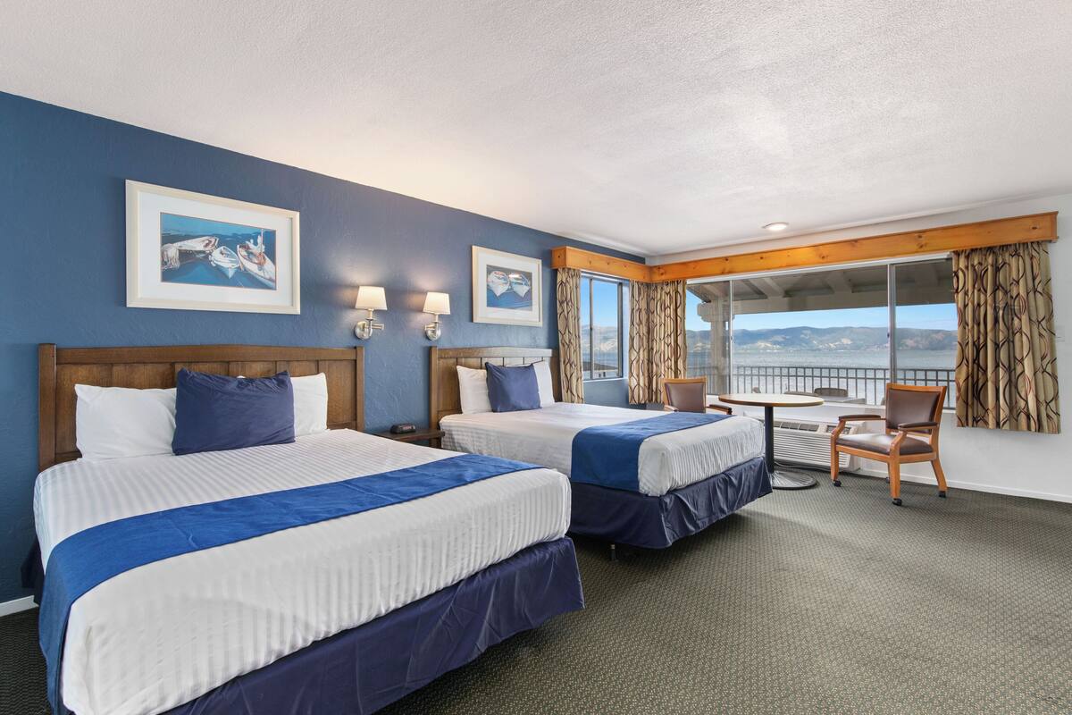 Habitación de hotel con dos camas, una mesita y una silla. Cuenta con dos ventanas con cortinas y un cuadro en la pared.