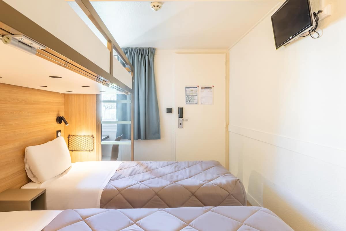 Ein Hotelzimmer mit einem hölzernen Kopfteil, einem Bett mit gesteppter Bettdecke, einem Nachttisch, einem Wand-Fernseher und einem Fenster mit Vorhängen.