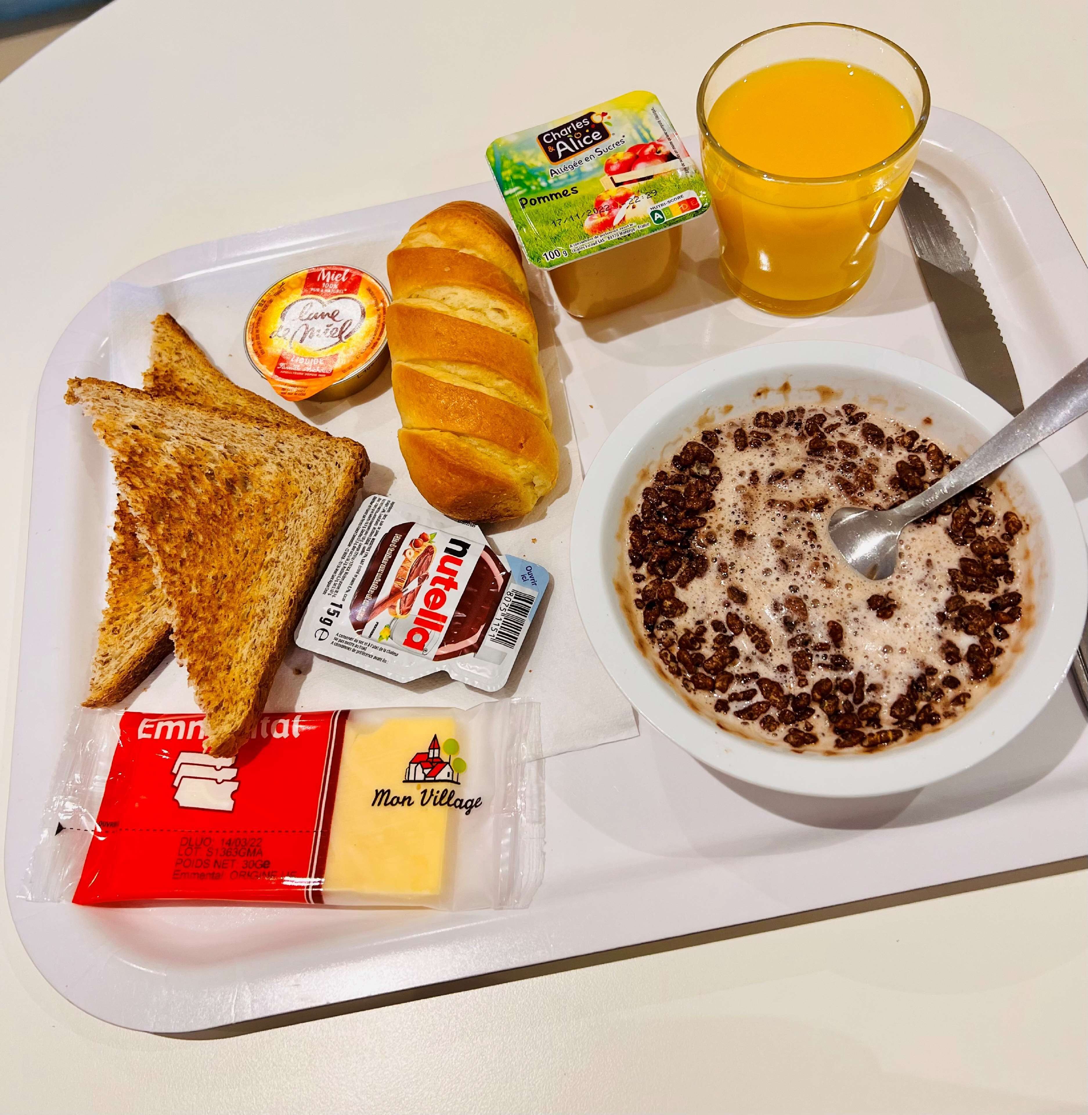 Una bandeja con tostadas, un croissant, un bol de cereal con leche, un vaso de zumo de naranja y una porción de mantequilla.