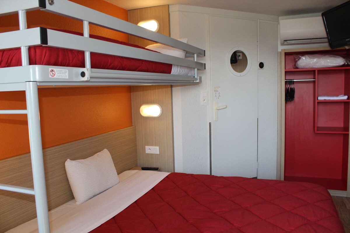 Una cama litera con colchón rojo, almohada blanca y manta roja. Se ve un armario blanco con espejo y un estante rojo con ropa.