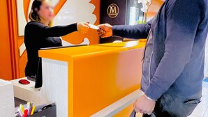 Un mostrador de recepción de hotel con una mujer detrás, un hombre con abrigo gris sosteniendo un bolso negro, y un cartel con la letra 'M' en la pared.
