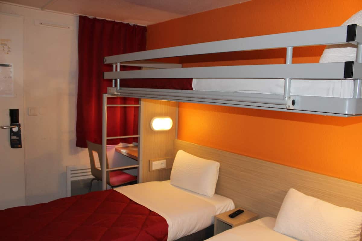 A hotel room with a bunk bed, a single bed, a bedside table with a lamp, and a red curtain.