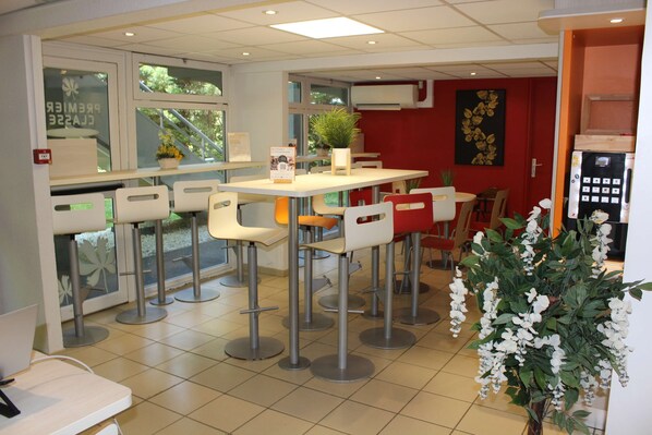 A restaurant with high tables and chairs, a potted plant, and a red wall with a floral painting. - Hotel Première Classe Saint Quentin En Yvelines - Elancourt (Elancourt)