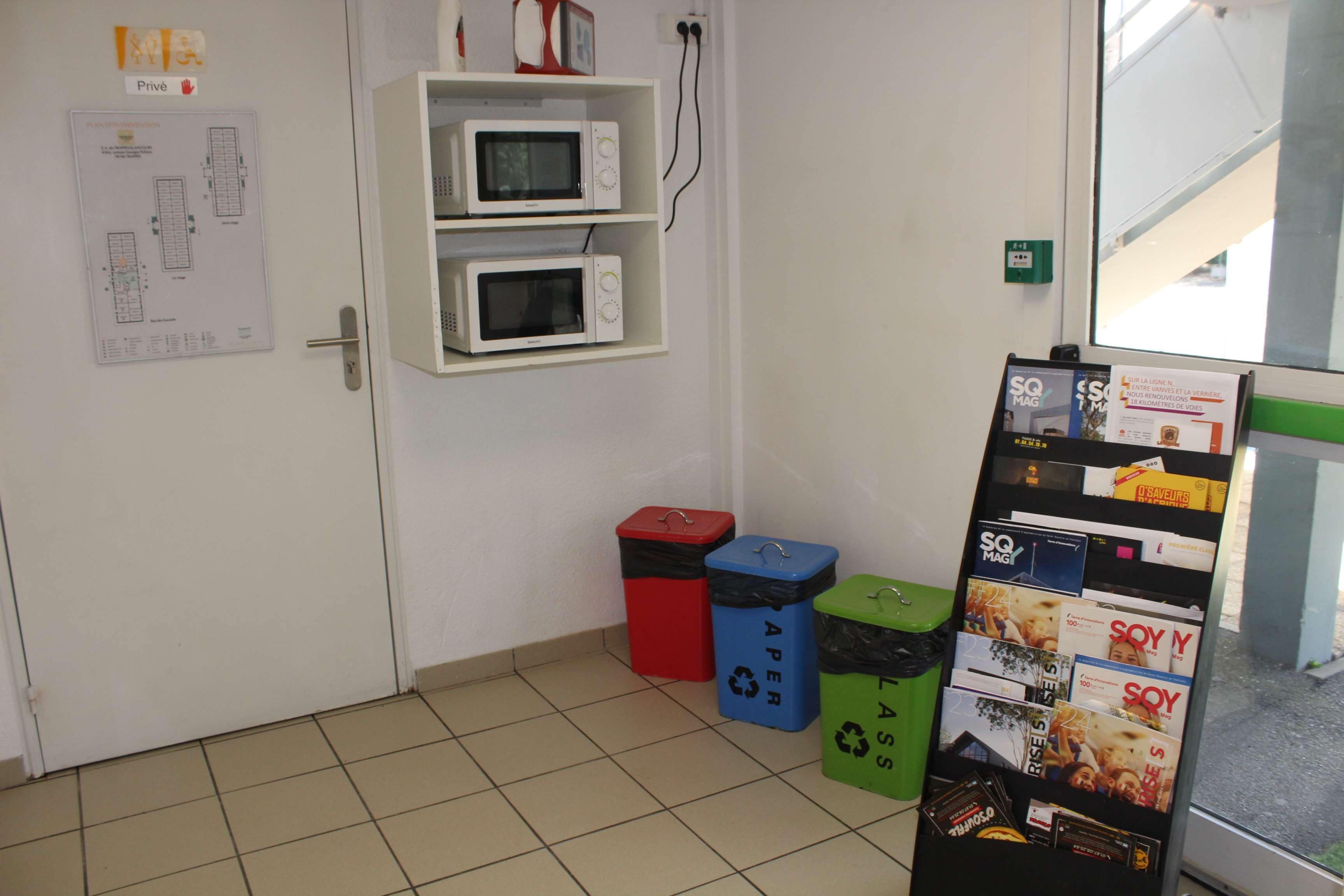 A room with a white door, two white wall-mounted microwaves, a recycling bin, and a display board with magazine covers.