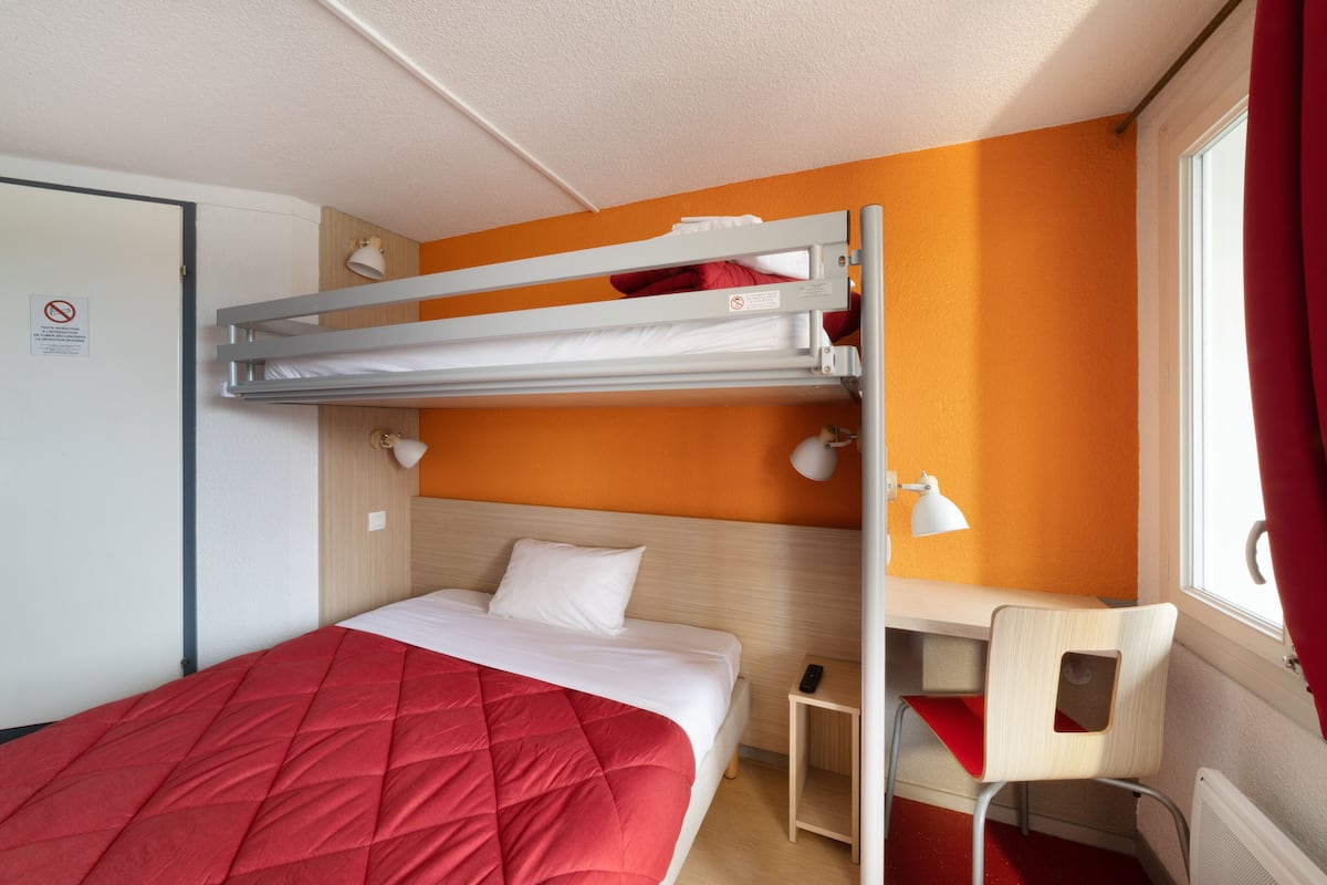 A bunk bed room with a red bedspread, a desk with a chair, and a window with curtains.