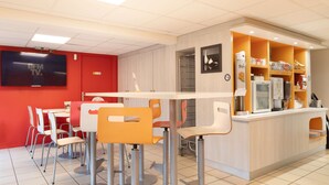 A modern cafe with a red accent wall, a TV screen, and a counter with various items. - Premiere Classe Meaux - Nanteuil les Meaux (Nanteuil-les-Meaux)