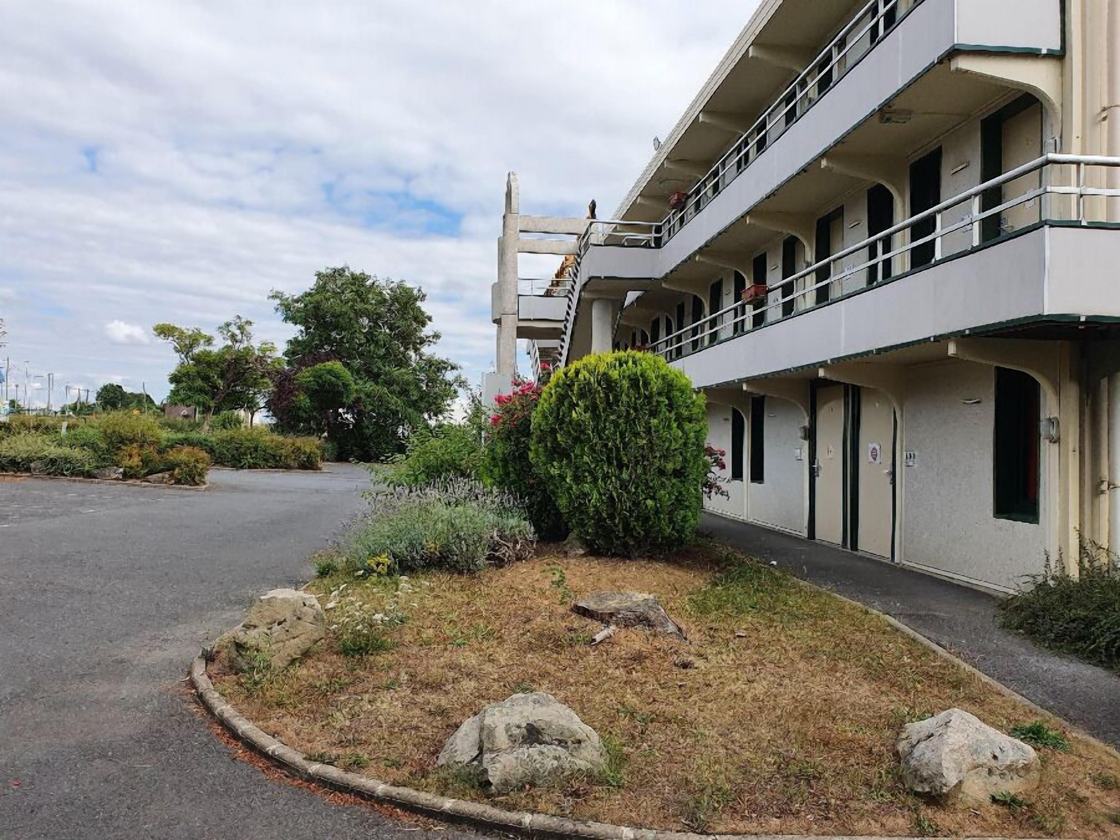 A building with a two-story balcony, a parking lot, and a landscaped area with shrubs and rocks.