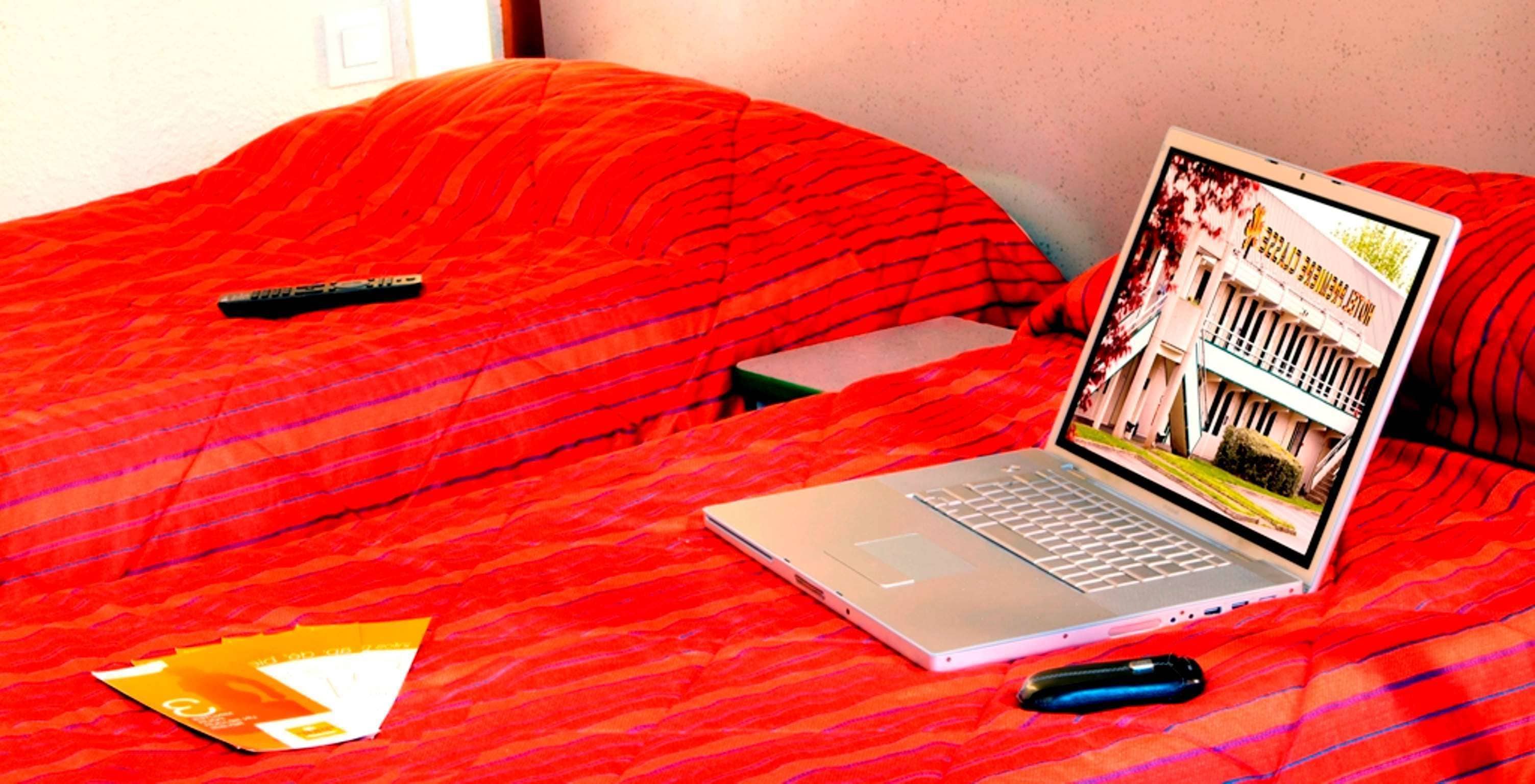 Un portatile su un letto con una coperta rossa, un telecomando e uno smartphone.