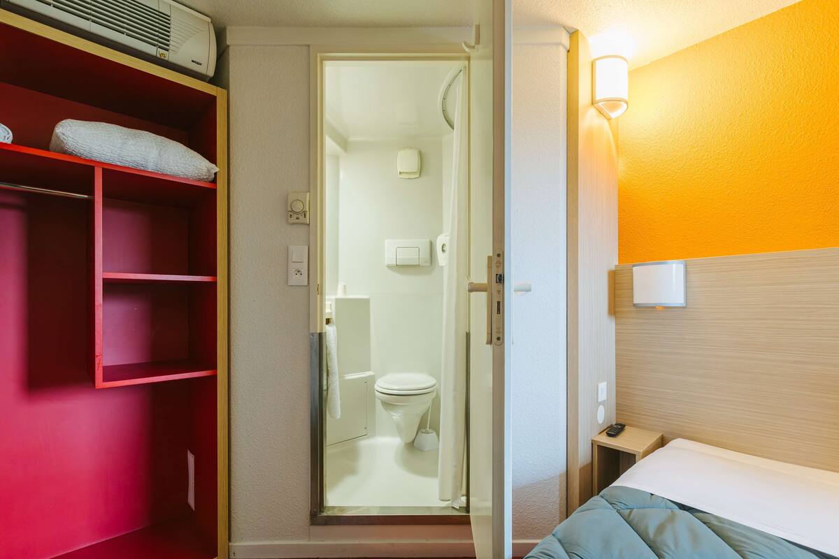 A hotel room with a bed, a shower area, and a red storage unit.