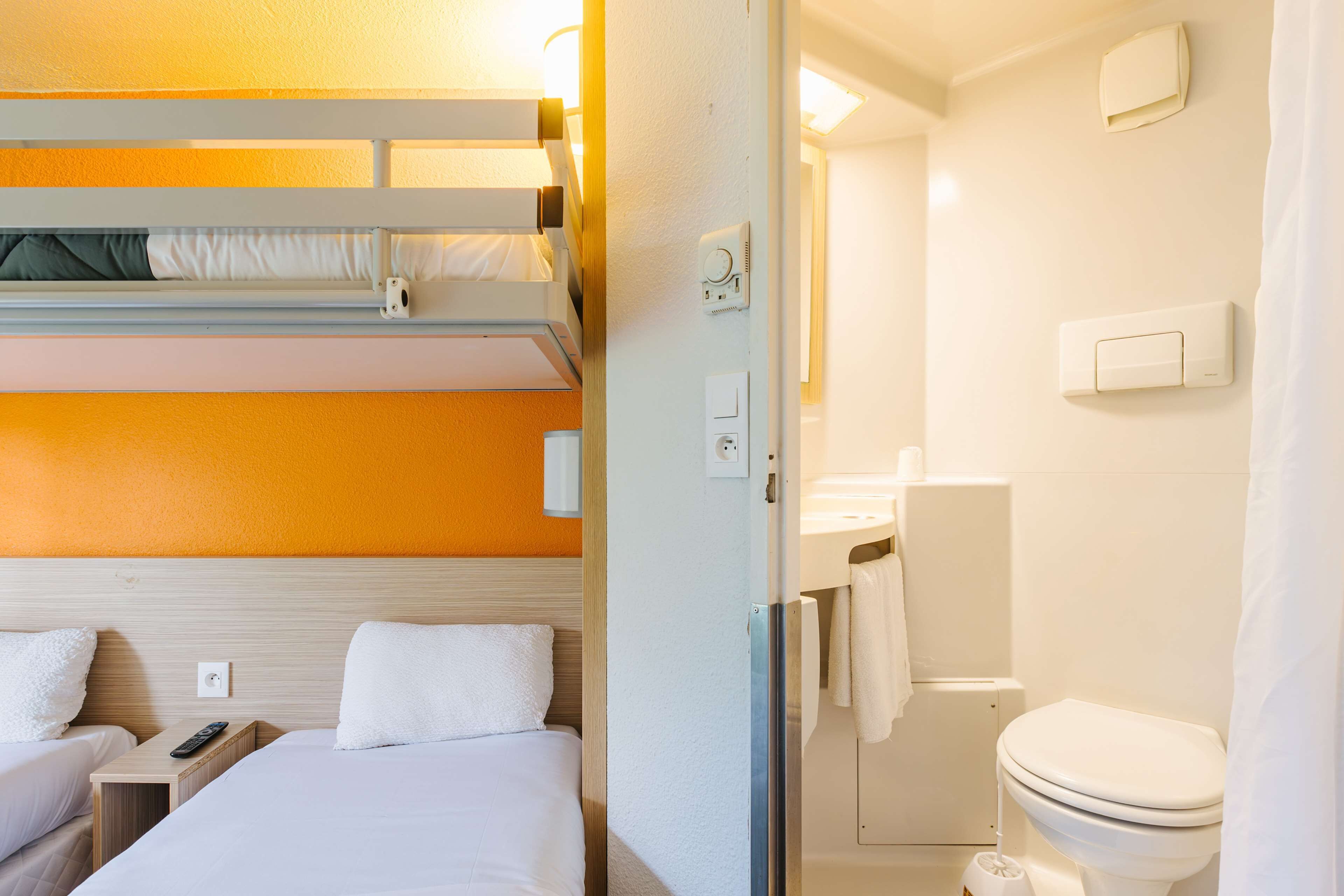 Una camera d'albergo con letti a castello, un letto singolo, un comodino con un telecomando, un bagno con un water, una doccia e un portasciugamani.