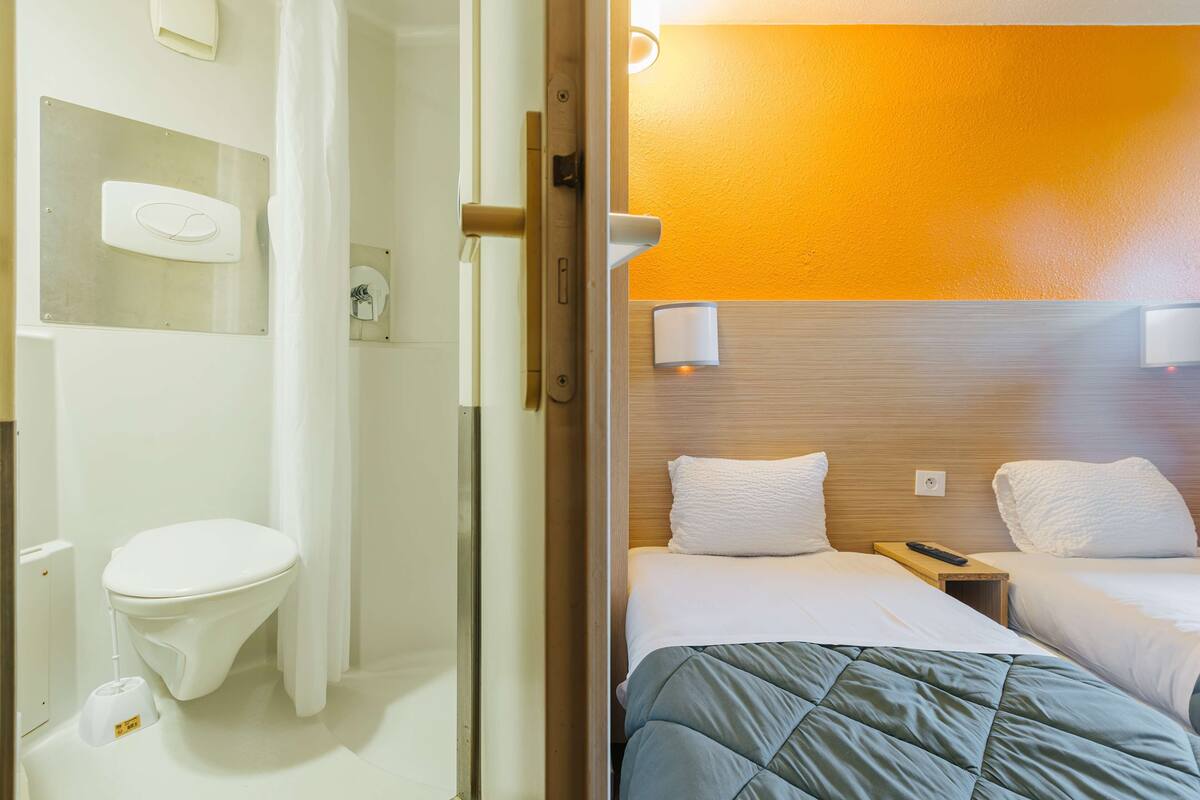 A hotel room with a wooden headboard, two beds with white bedding, a bedside table with a remote control, and a white toilet with a bidet attachment.