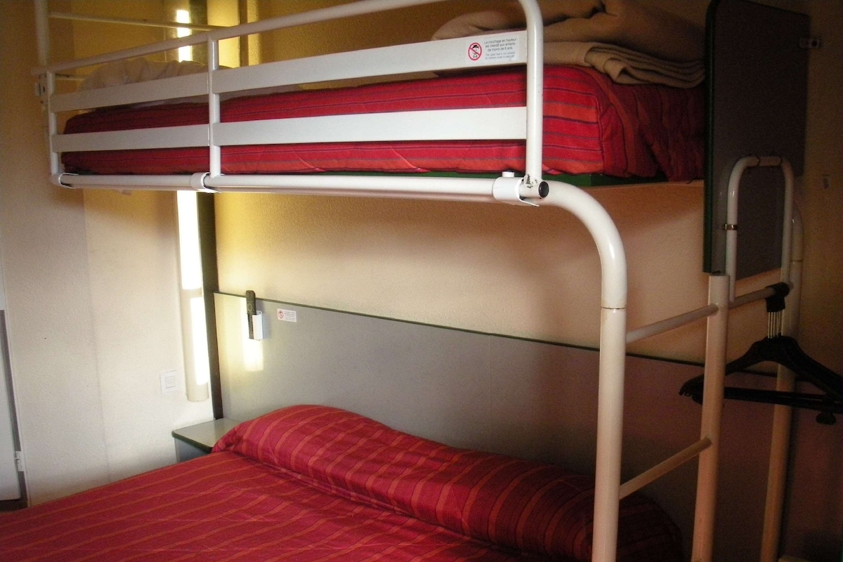 Un lit superposé avec un matelas rouge et un lit inférieur avec un oreiller rouge. Il y a un mur blanc avec un interrupteur et une petite table avec une lampe.
