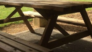 Ein Picknicktisch mit robuster Holzkonstruktion und Bänken, auf einer Betonfläche, mit Rasen und einem Gebäude im Hintergrund.