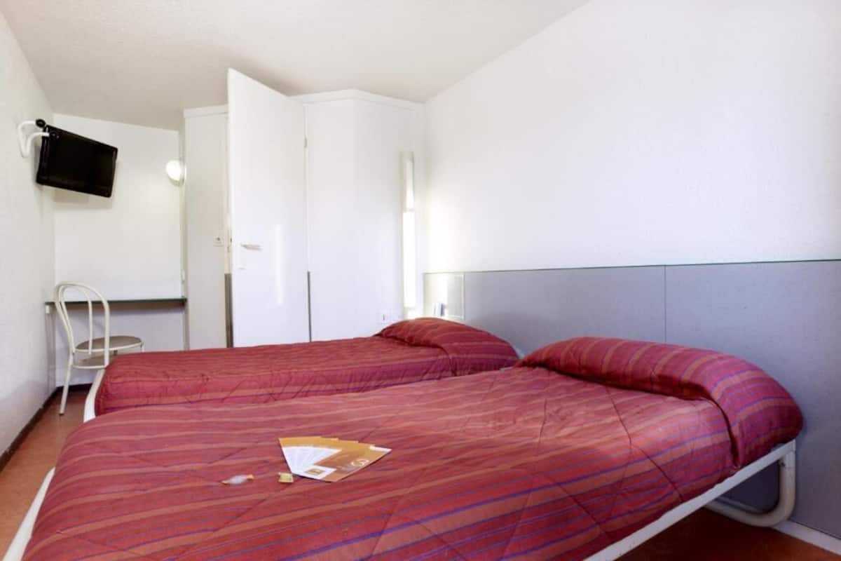Une chambre avec un lit simple, une couvre-lit à rayures rouges, un téléviseur à écran plat fixé au mur et une armoire blanche.