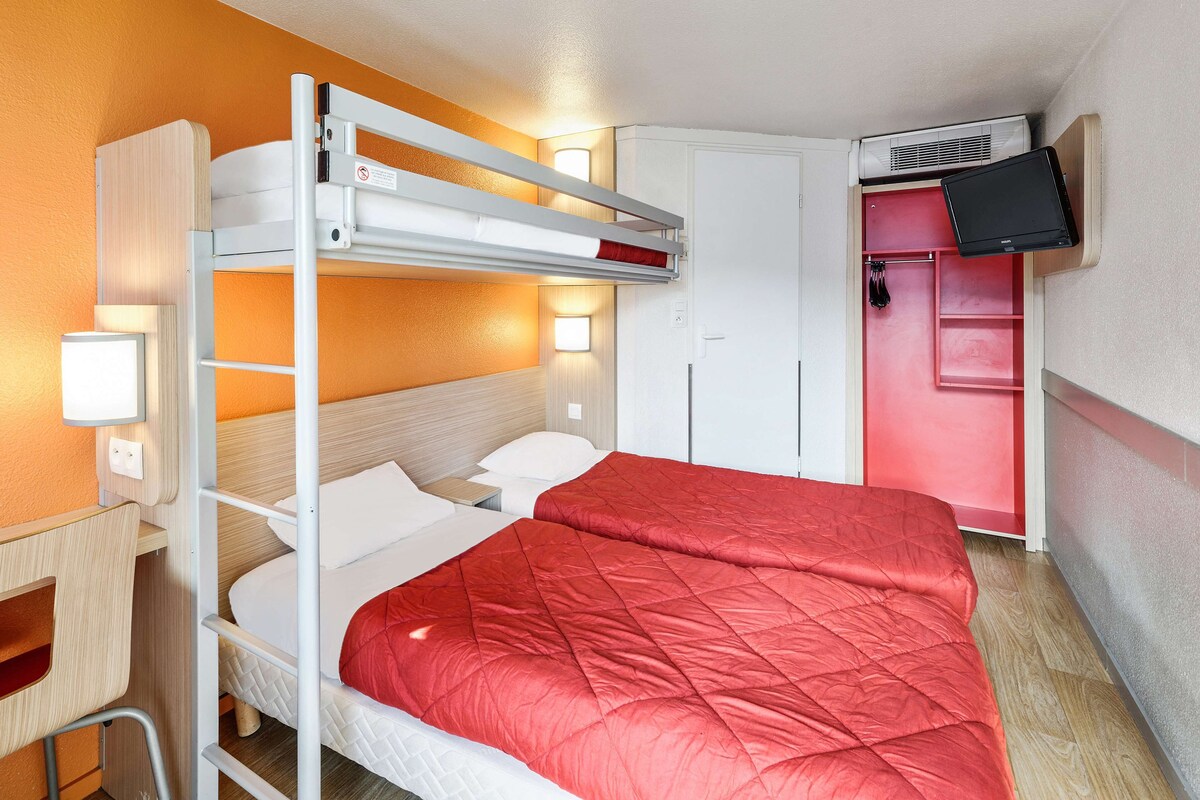 Un lit superposé avec une couverture matelassée rouge, un bureau avec une chaise, une armoire et un téléviseur fixé au mur.
