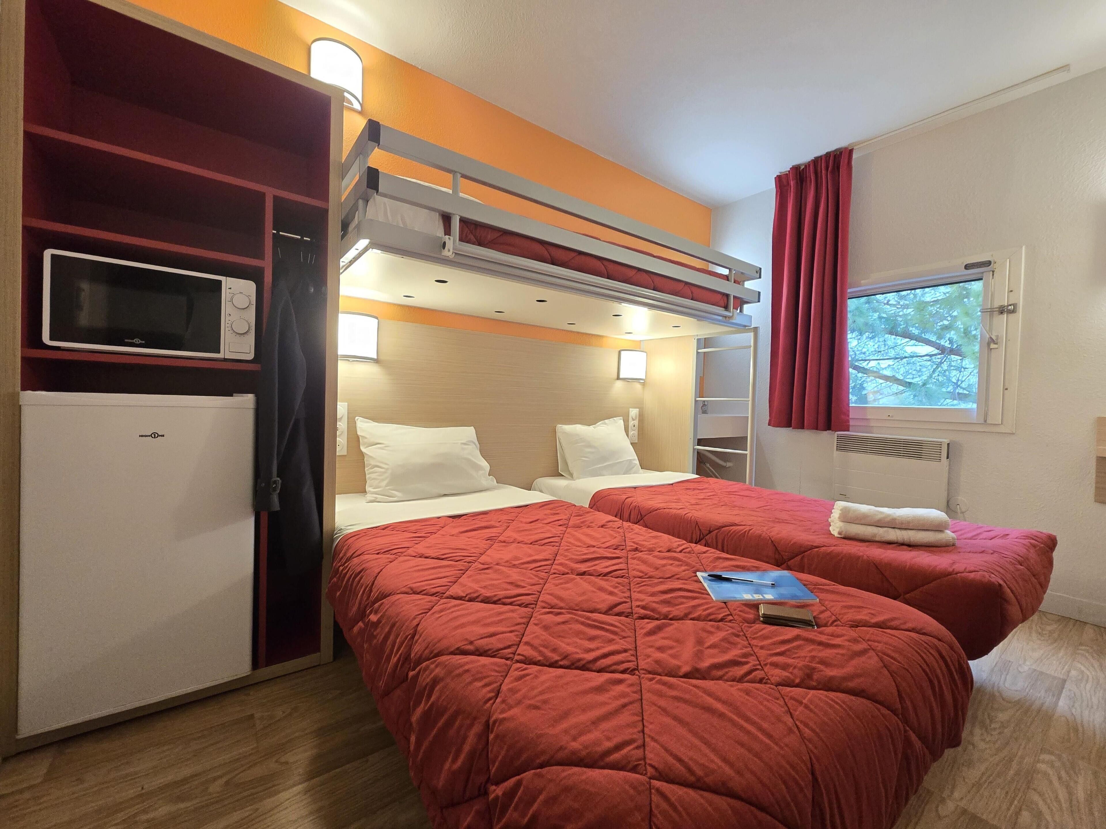 A room with two beds, a microwave, a mini-fridge, a window with curtains, and a shelf with a TV.