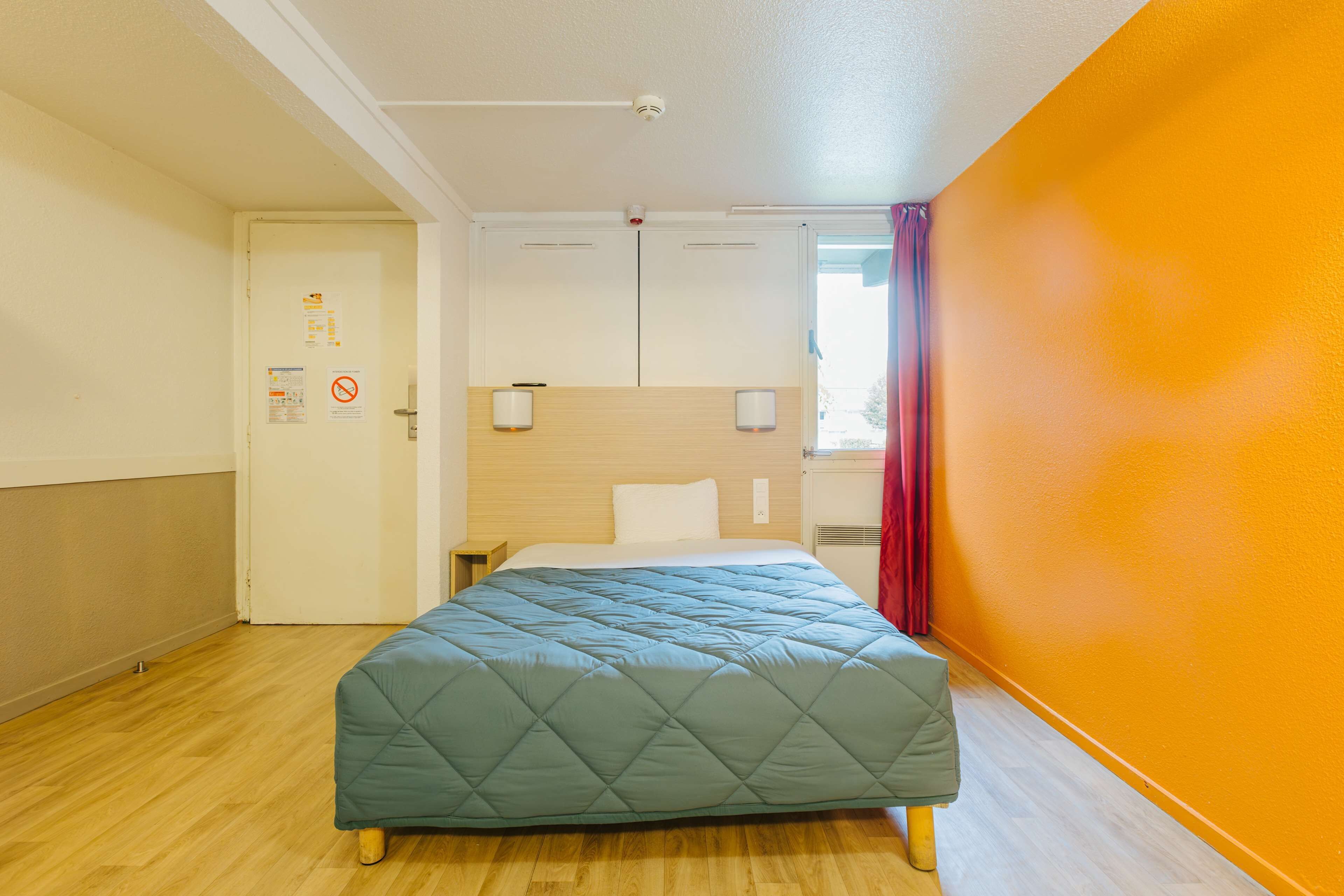 Een hotelkamer met een bed, houten vloer, een deur met een bordje en een oranje wand.