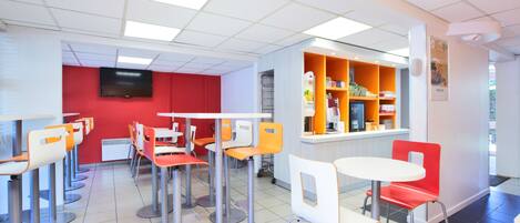 Una cafetería luminosa con varias mesas y sillas, una pared roja con un televisor y estantes con productos alimenticios.