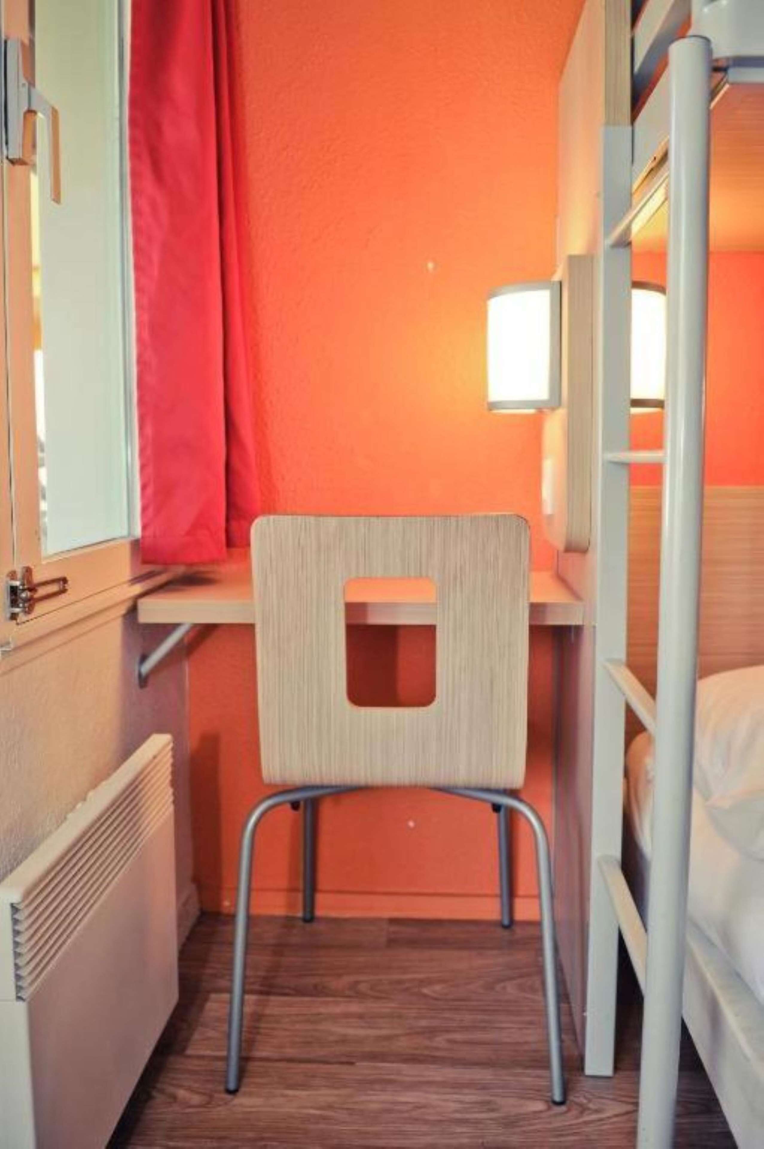 A compact room with a bunk bed, a small desk with a chair, a window with a curtain, and a radiator.