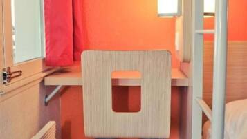 A compact room with a bunk bed, a small desk with a chair, a window with a curtain, and a radiator.