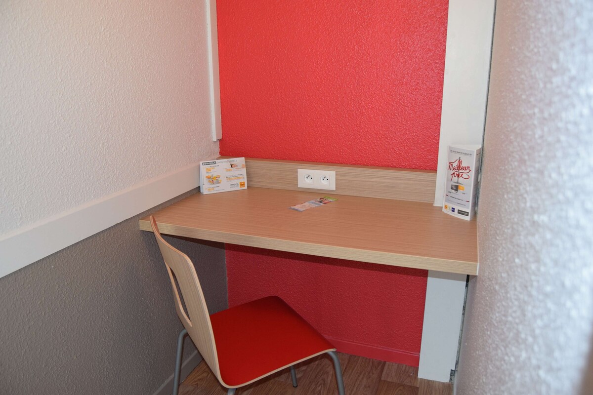 Una oficina pequeña y privada con un escritorio de madera, una silla roja y una pared decorada en rojo. Hay dos enchufes en la pared, y una caja con un logotipo en el escritorio.