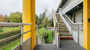 Un escalier mène à un bâtiment dont la façade est jaune, orné d’une rambarde métallique, et qui offre une vue sur un paysage verdoyant avec des arbres et une route.