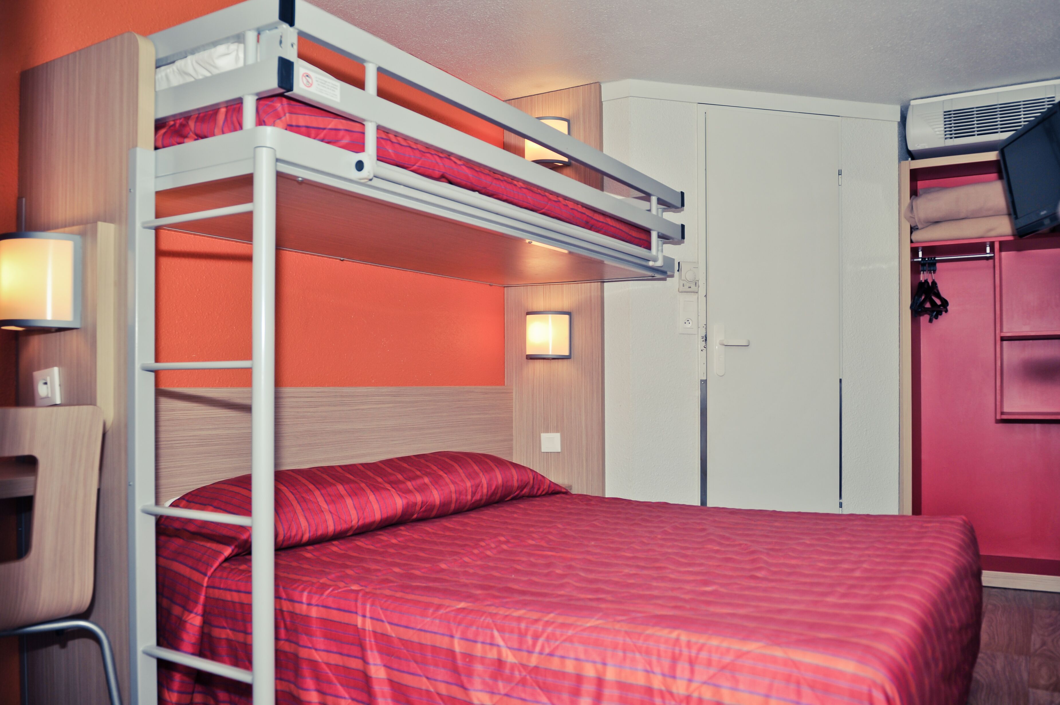 Una cama litera con colcha roja, un escritorio con silla y un armario con espejo.