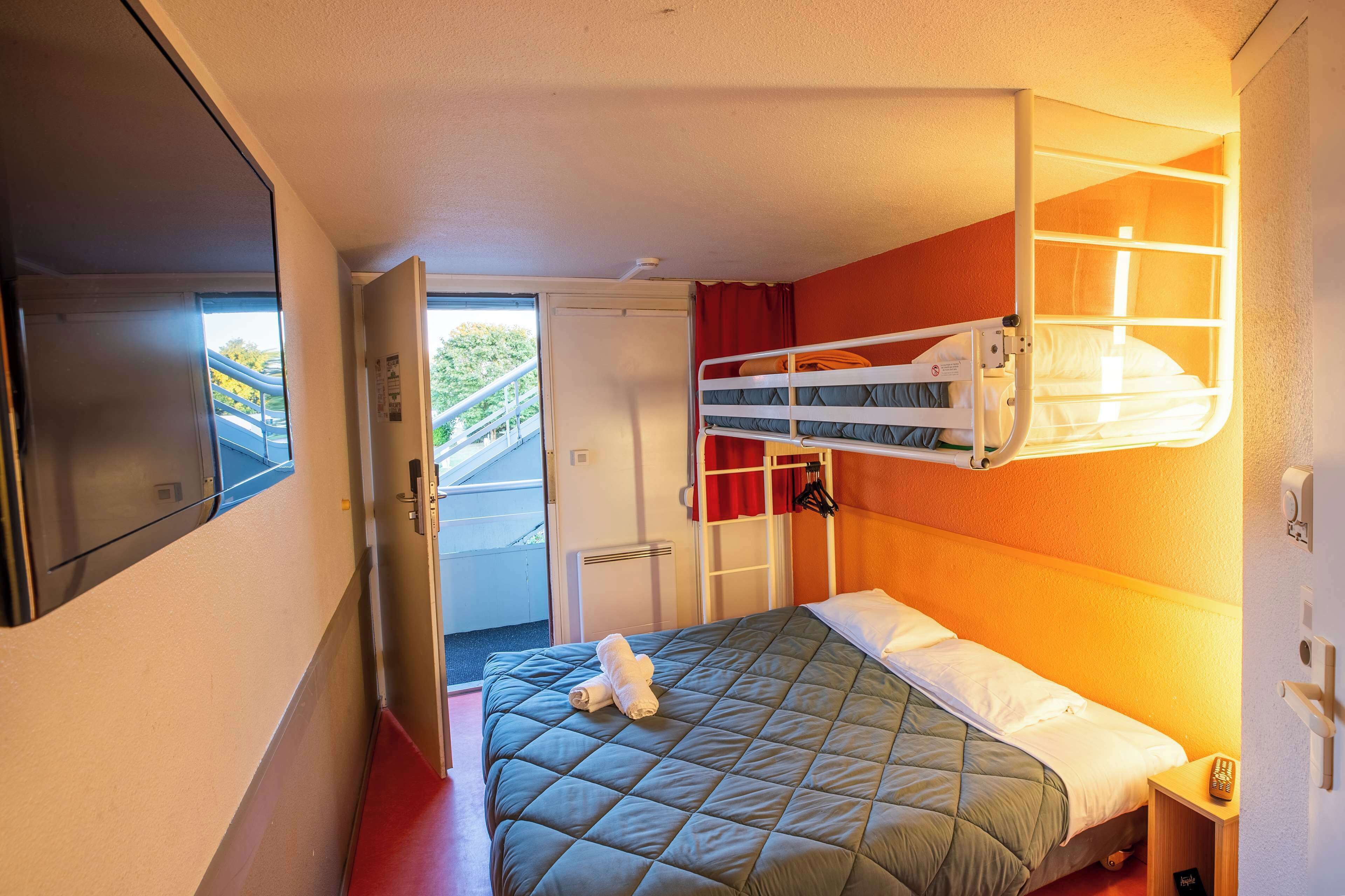 A compact hotel room with a bunk bed, a single bed, a small bedside table, a window, and a door leading to a balcony.
