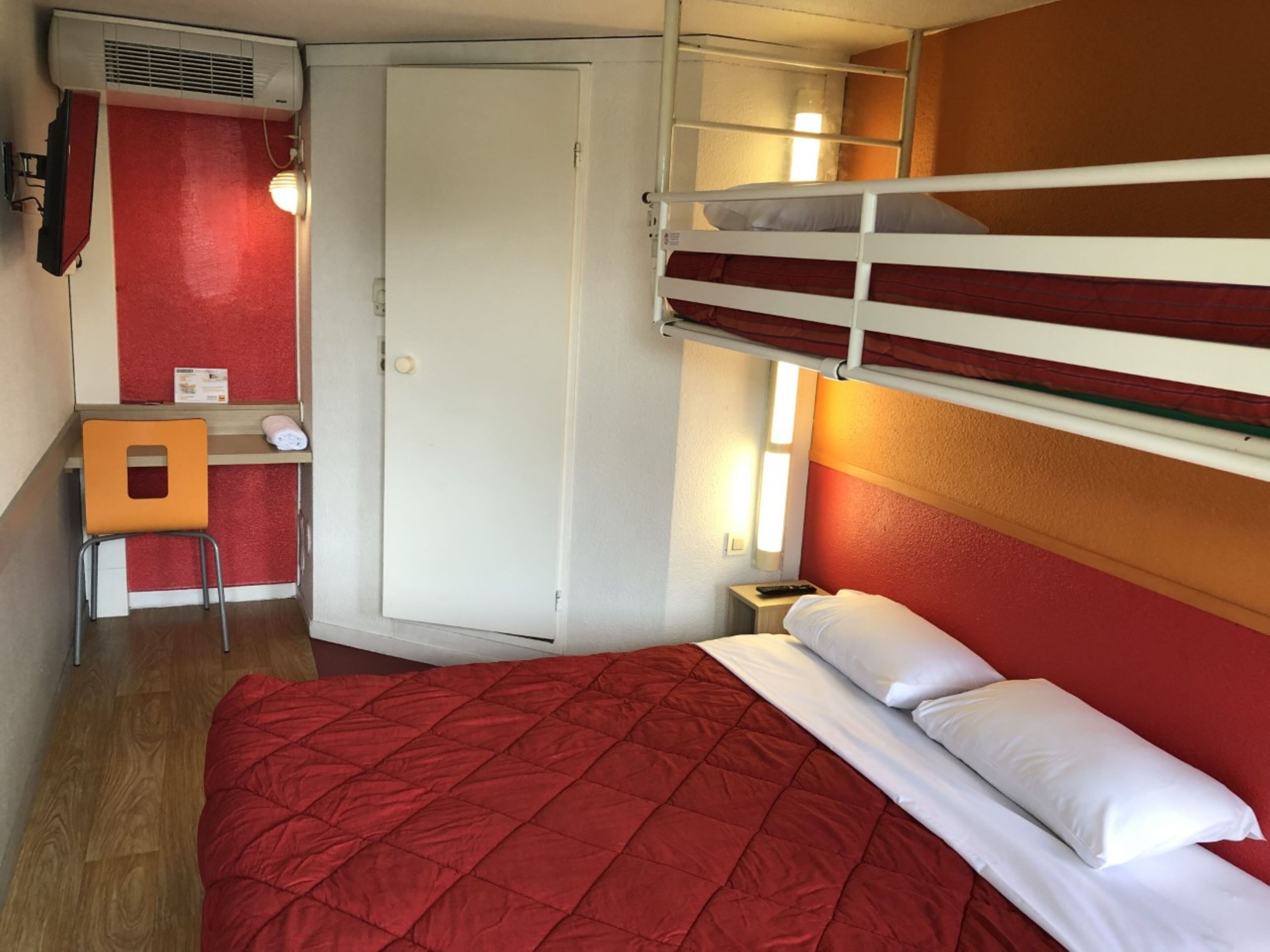 Ein kleines Hotelzimmer mit Etagenbett, Schreibtisch und Stuhl, Kleiderschrank und Nachttisch mit Lampe.