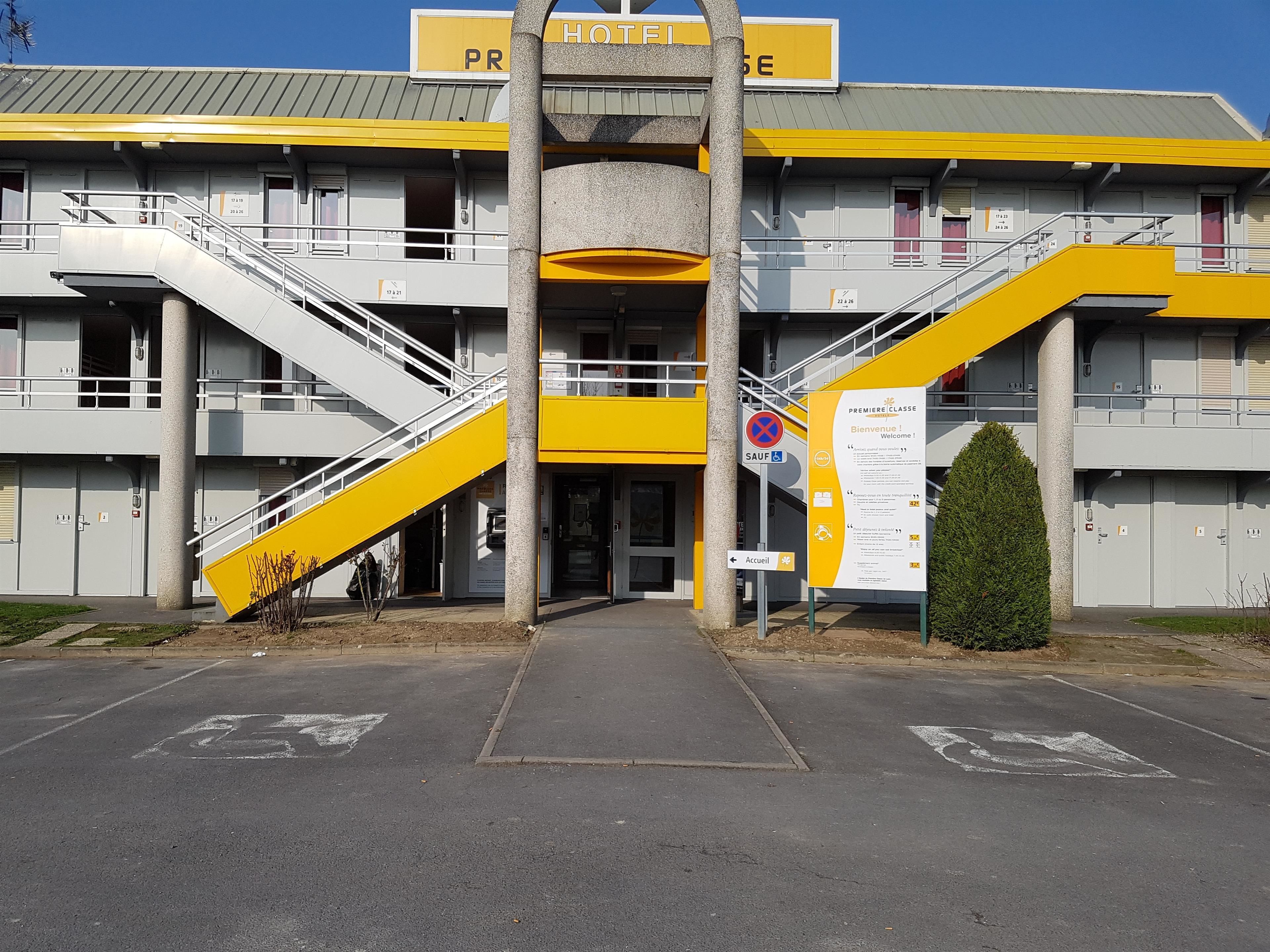 A hotel entrance with a yellow and grey facade, multiple staircases, and a sign indicating a no parking zone.