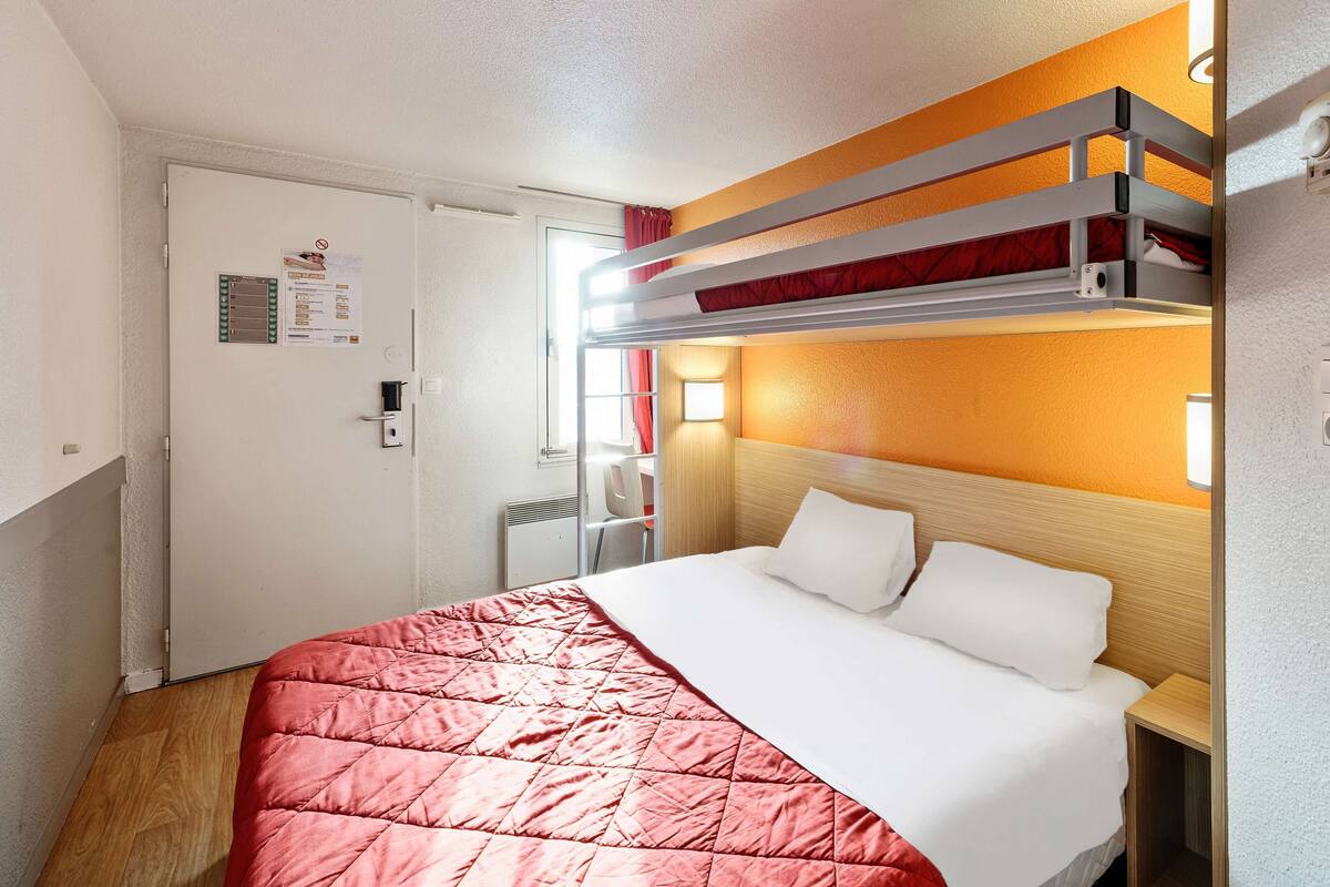 A hotel room with a bunk bed, a single bed, a wooden headboard, a red quilted bedspread, a white pillow, a bedside table, and a door with a notice.