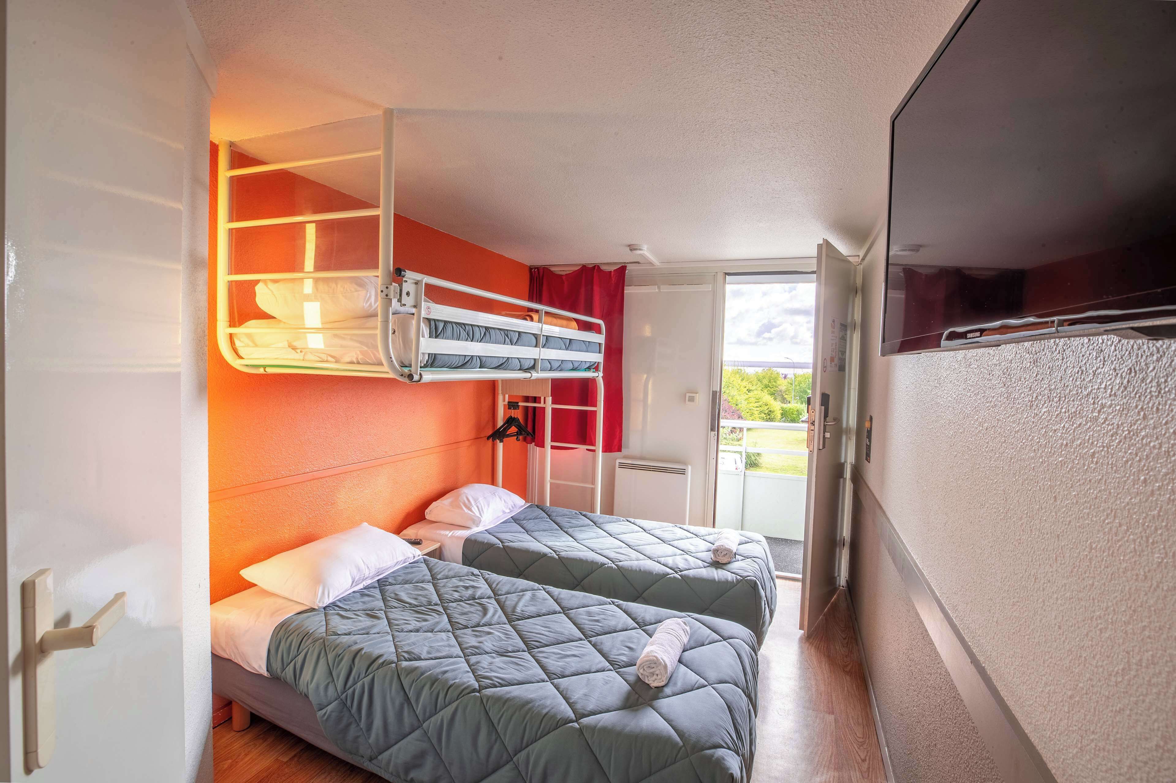 Kompakti hotellihuone, jossa on kaksi sänkyä, kerrossänky, liukuovi parvekkeelle ja suuri ikkuna.