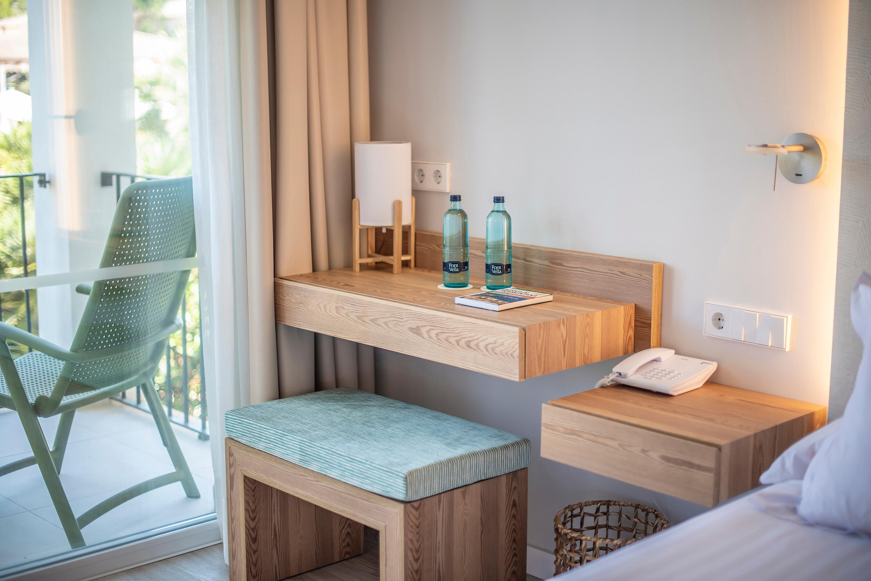 A hotel room with a wooden desk, a green chair, a small bench, a bedside table with a phone, and two bottles of water.