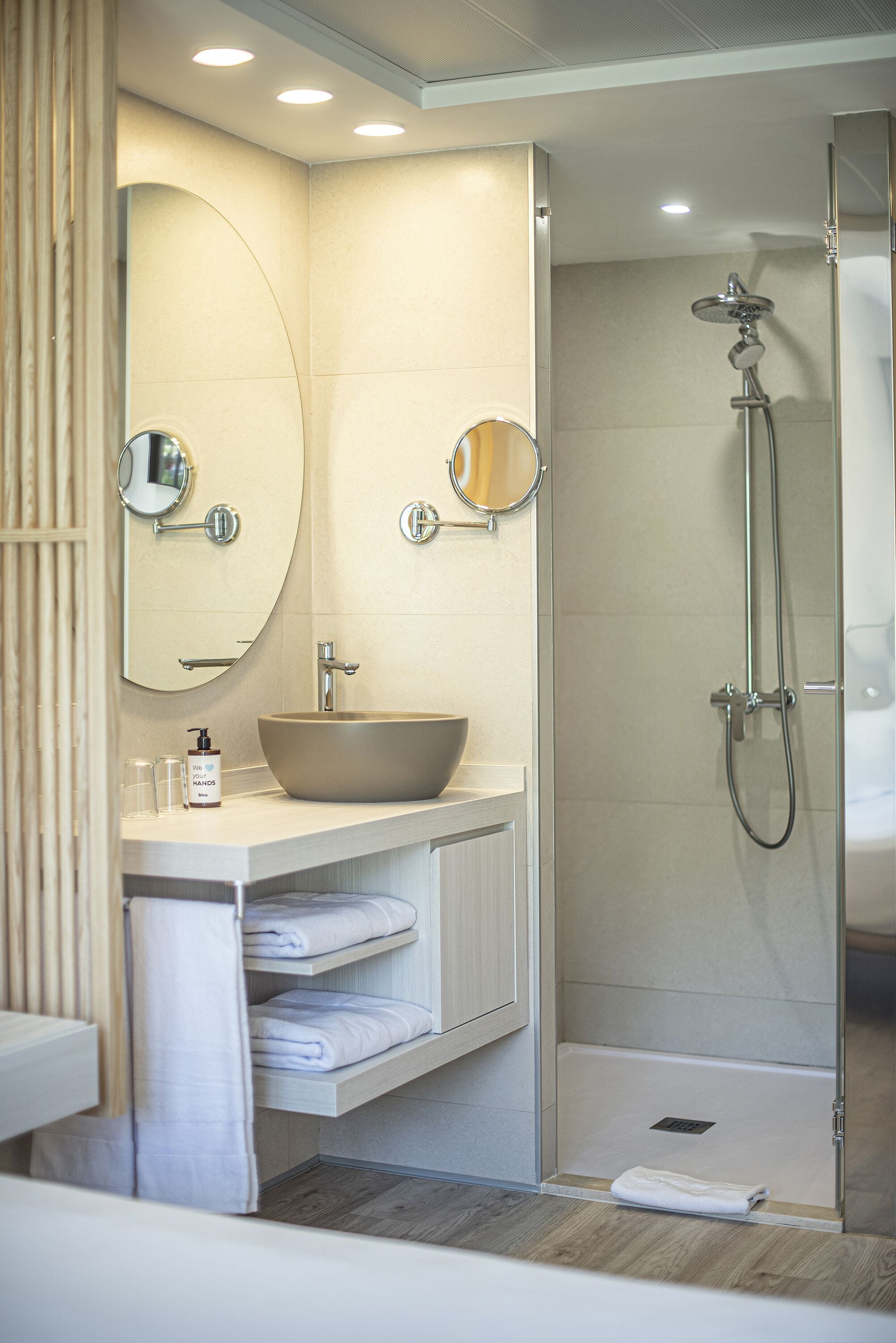 Une salle de bain moderne avec un grand lavabo sur pied, deux miroirs fixés au mur, une douche à parois de verre et des serviettes soigneusement pliées, rangées sur une étagère.