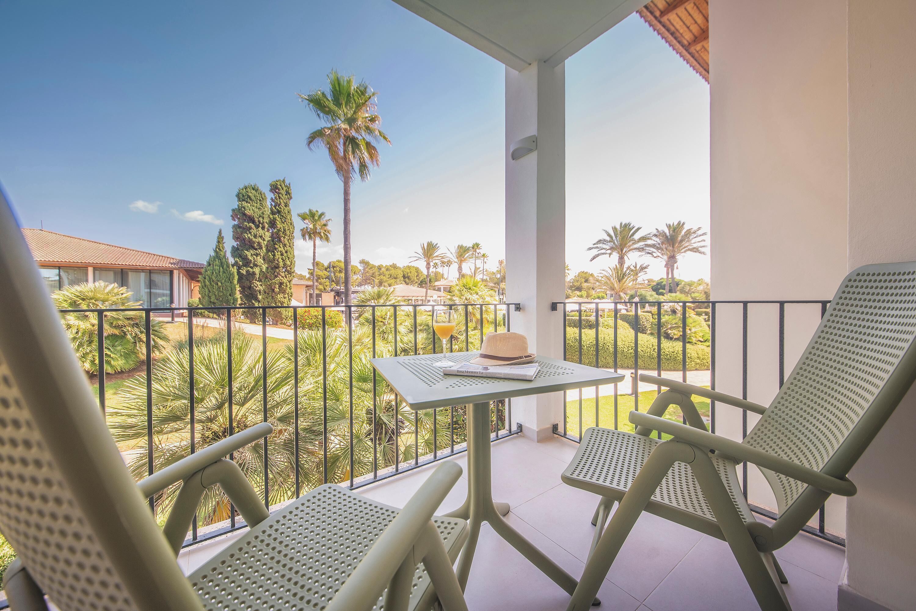 Un balcon avec deux chaises vertes et une table, donnant sur un jardin avec des palmiers et un bâtiment en arrière-plan.
