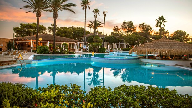 A swimming pool with a thatched-roof structure, palm trees, and a building with a tiled roof. - Blau Colònia Sant Jordi (Ses Salines)