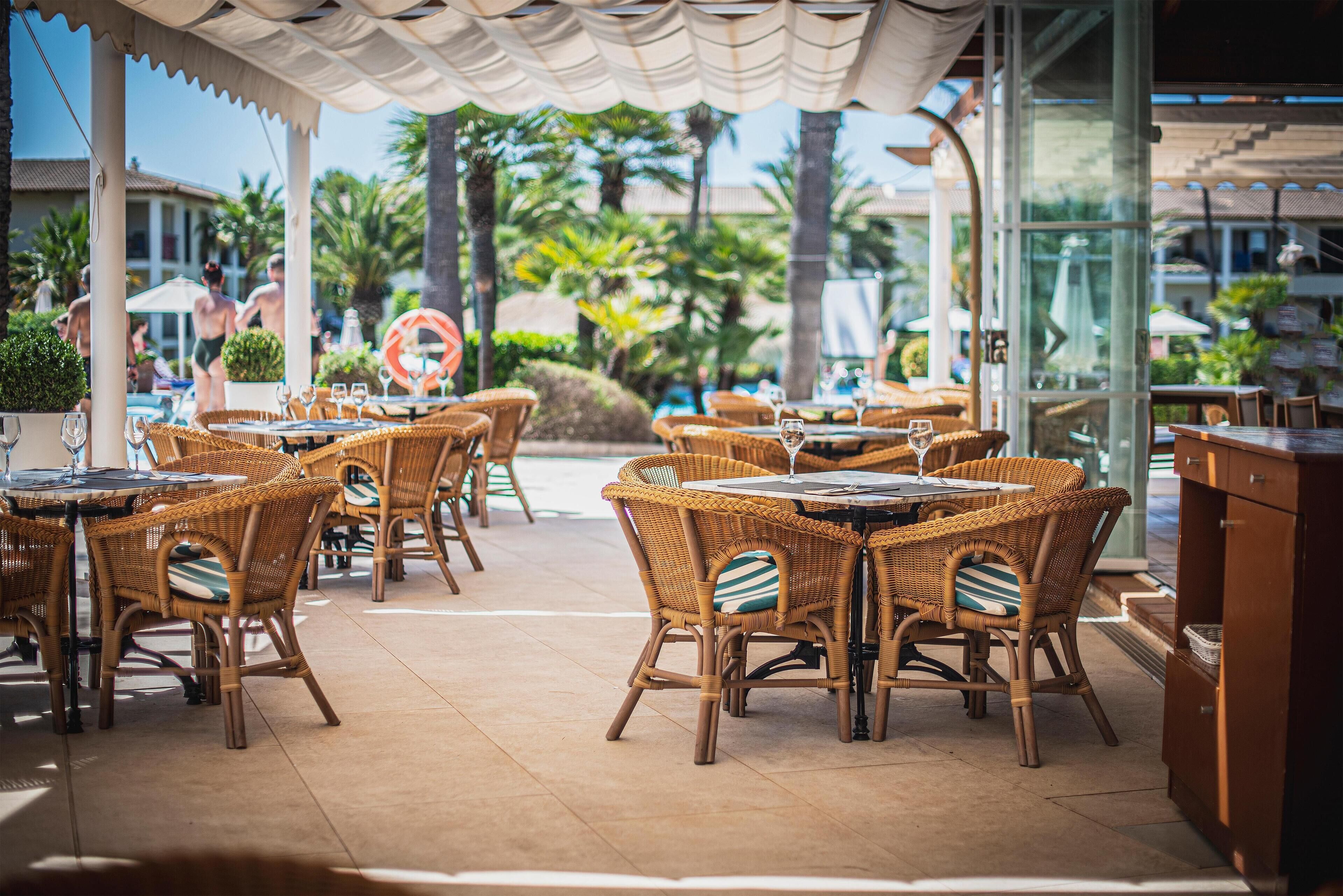 An outdoor dining area with wicker tables and chairs, a pool in the background, and a tropical garden setting.