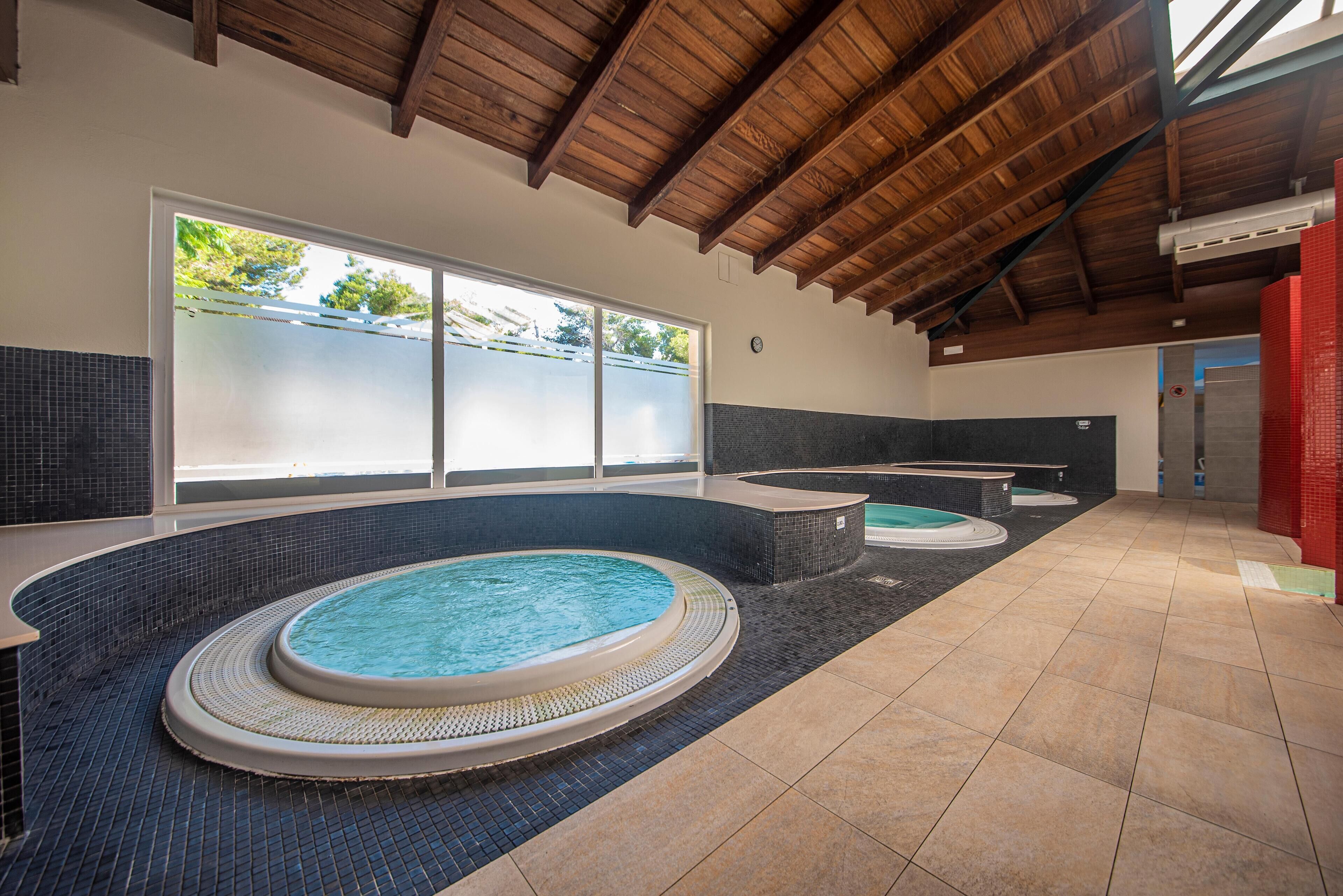 Una amplia piscina interior con varios jacuzzis, suelo de baldosas y techo de madera.