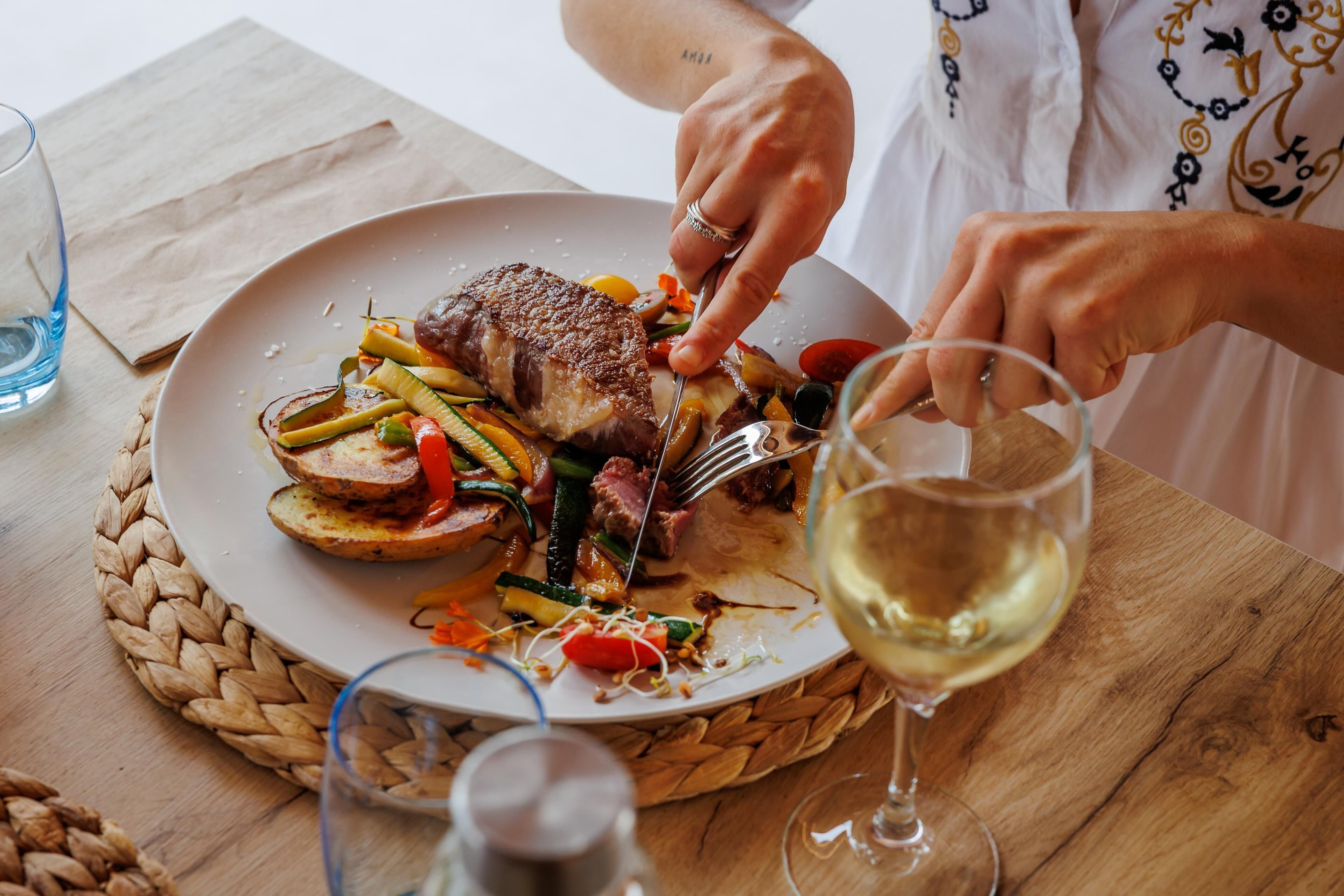 A person dining at a table with a plate of food, a glass of white wine, and a woven placemat.