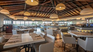 A spacious restaurant with high ceilings, large pendant lights, and a central bar area. Tables are neatly arranged with place settings. - Blau Colònia Sant Jordi (Ses Salines)
