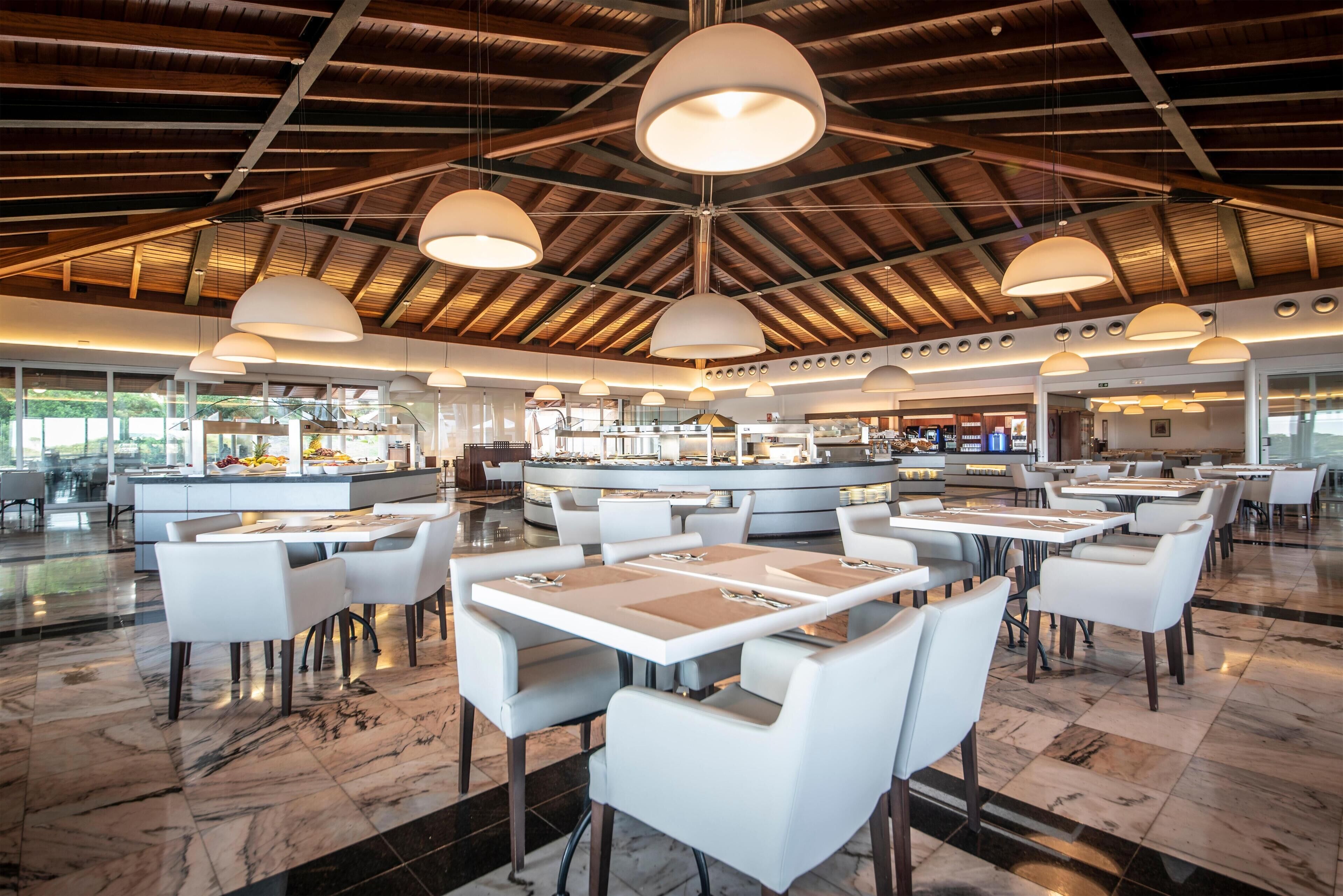 A spacious restaurant with high ceilings and modern pendant lighting. Tables are arranged in a U-shape around a central island with a kitchen.