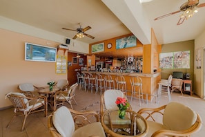 A bar area with multiple seating options, including wicker chairs and bar stools, and a television screen mounted on the wall.