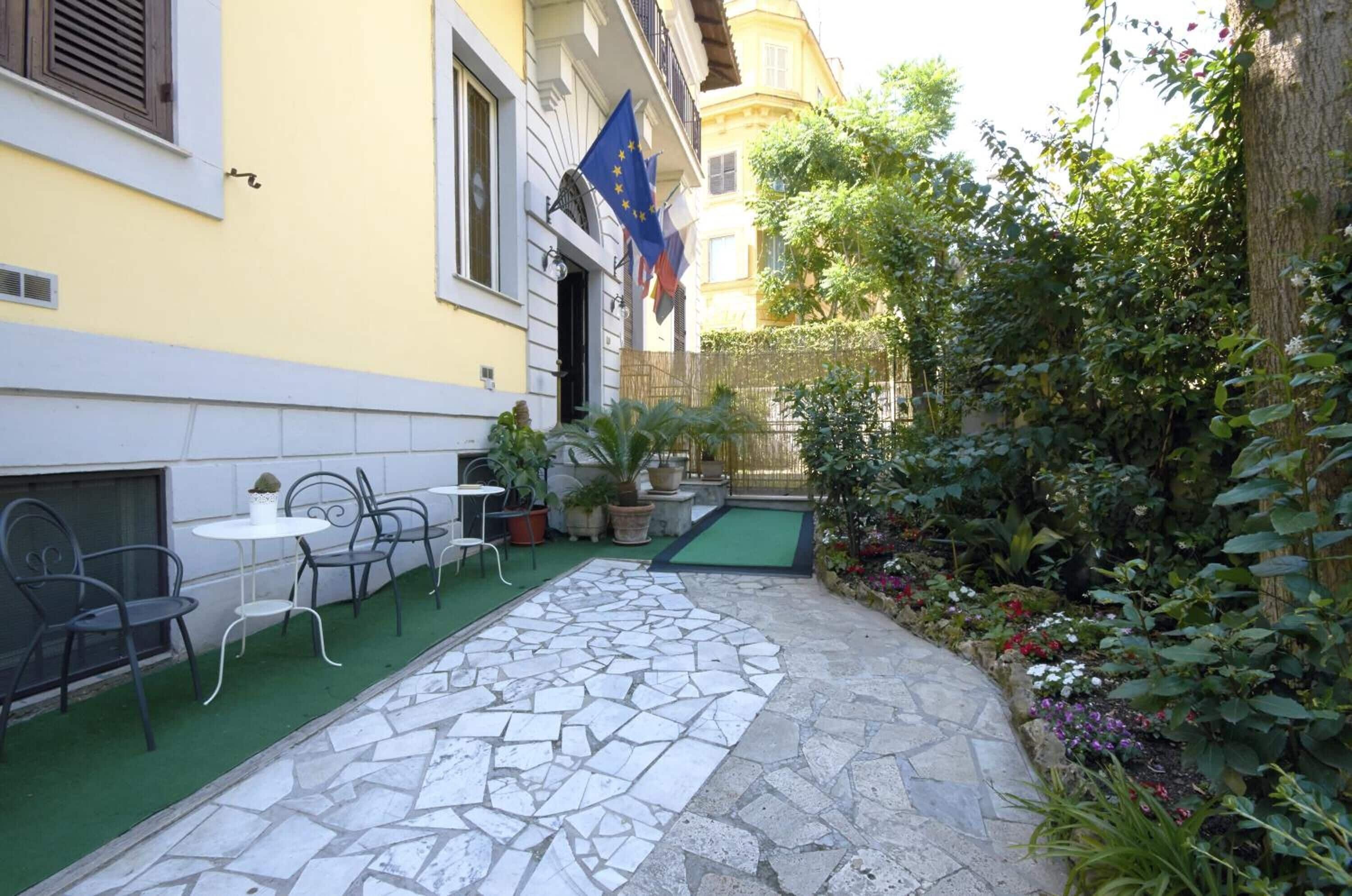 Un patio con mesas al aire libre, un sendero de piedra y diversas plantas y flores.
