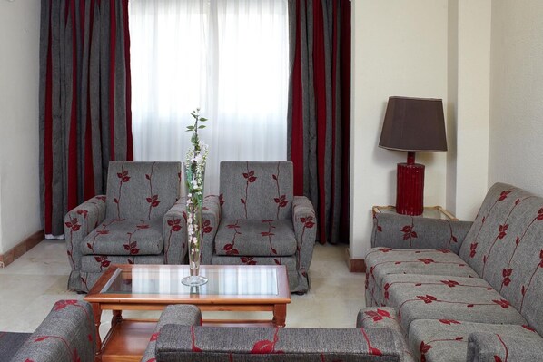 Un salon avec deux canapés gris ornés de motifs floraux rouges, une table basse en verre, une lampe rouge et un vase contenant des fleurs blanches.
