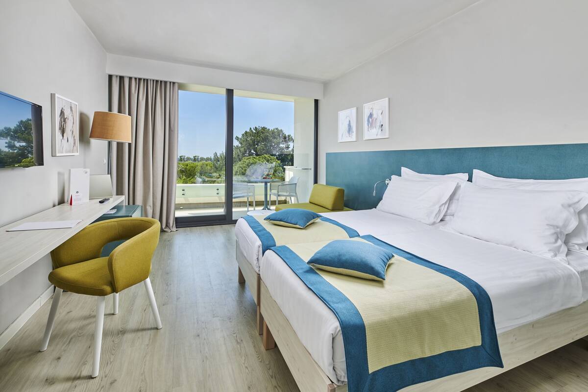 A hotel room with a bed, two bedside tables, a desk, a chair, and a view of the outdoors.