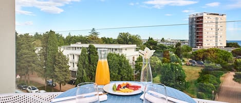A balcony table with a plate of fruit, a glass of orange juice, and a vase of flowers, overlooking a residential area with trees and buildings.