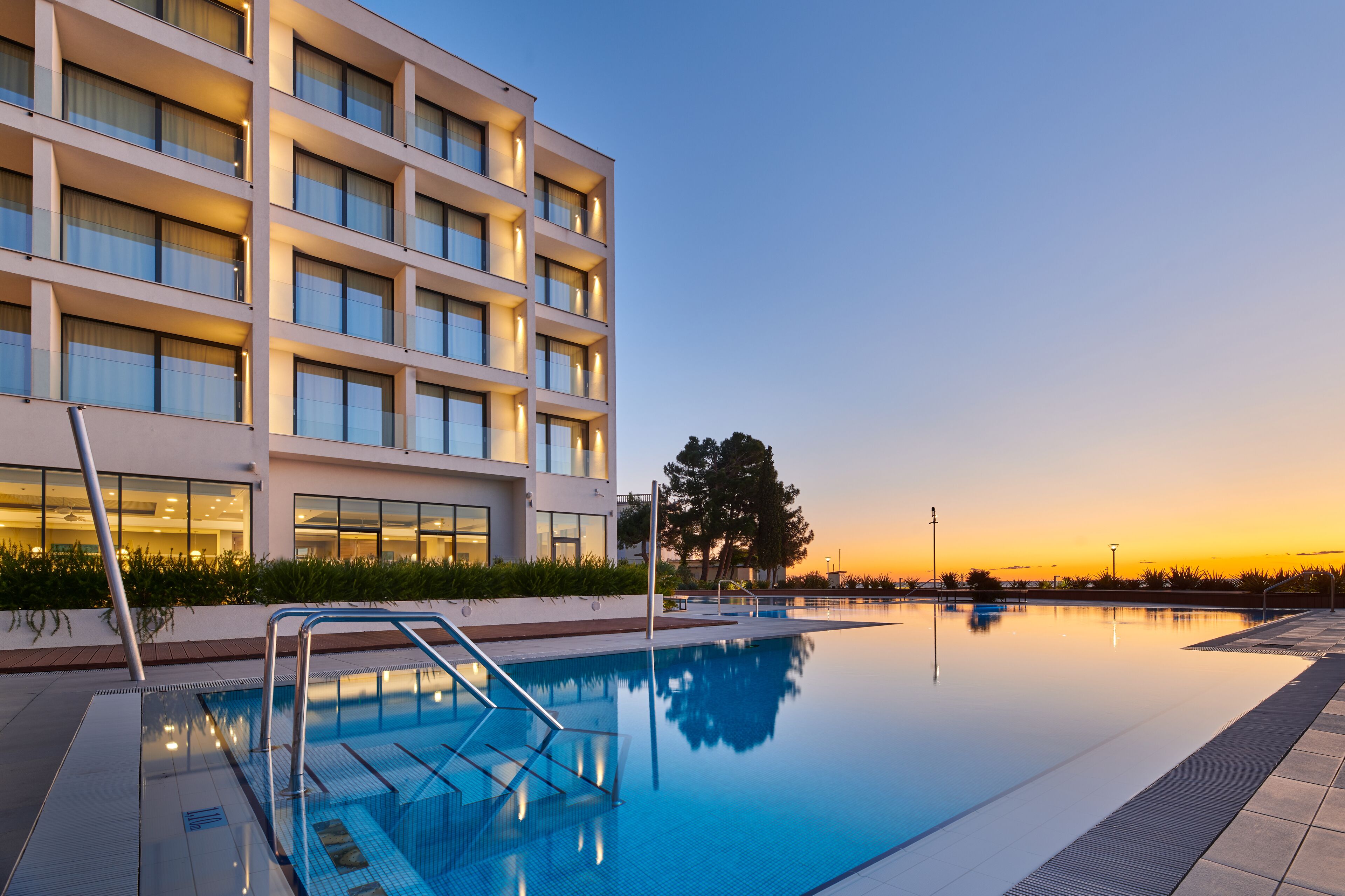 A modern hotel with a swimming pool, large windows, and a view of the sunset.